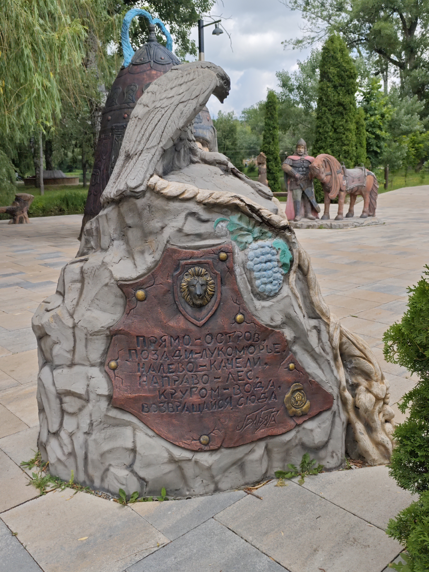 The image displays an outdoor sculpture park or memorial in Cherkessk, Russia. The central focus is a large, stylized rock sculpture adorned with a plaque. The plaque features Cyrillic text, a depiction of a lion's head in a shield, and a cluster of grapes. To the left, a large, dark bell-shaped structure with a blue rope detail stands partially visible. To the right of the main sculpture, a statue of a warrior in armor, with a red cape, stands beside a statue of a horse. In the background, there are trees, other sculptures, and a paved walkway. The environment appears to be a park or public garden, with greenery and stone paving. The sky is partly cloudy.