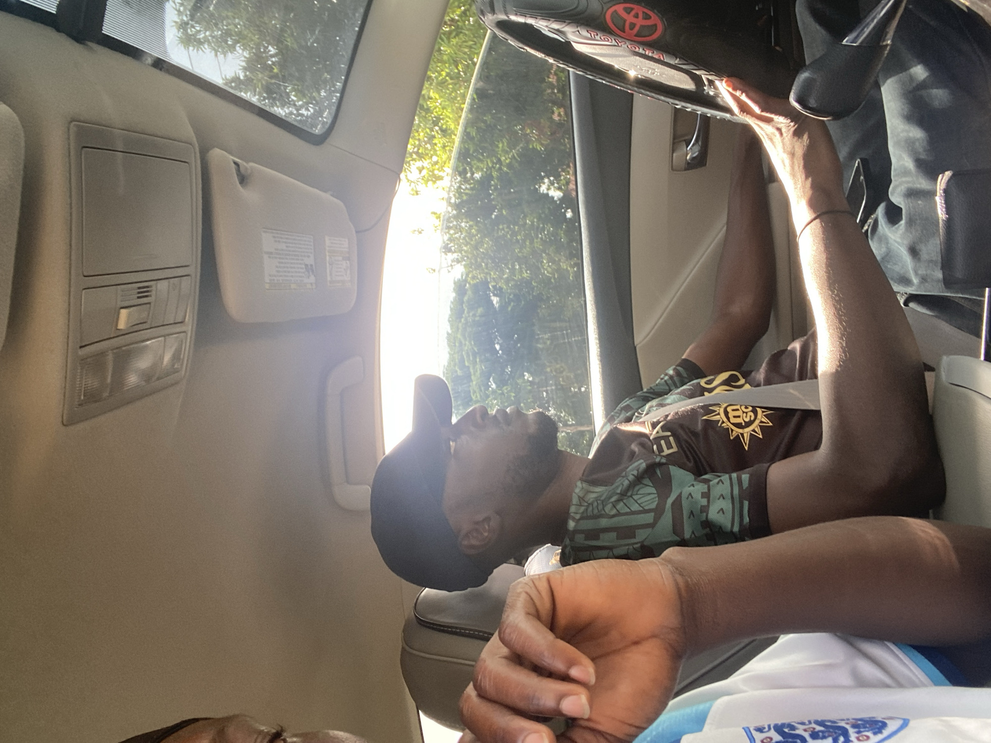 Two individuals are inside a vehicle. The person in the foreground is partially visible on the left, wearing a white shirt with blue accents. The driver, wearing a black baseball cap and a green and brown patterned shirt, is seated in the driver's seat, holding the steering wheel. The steering wheel has a red Toyota logo. The scene is illuminated by sunlight streaming through the car windows, casting shadows. Trees and foliage are visible outside the car. The interior of the car appears to be a standard passenger vehicle. The photograph was taken in Bar Jirgi Summa, Nigeria.