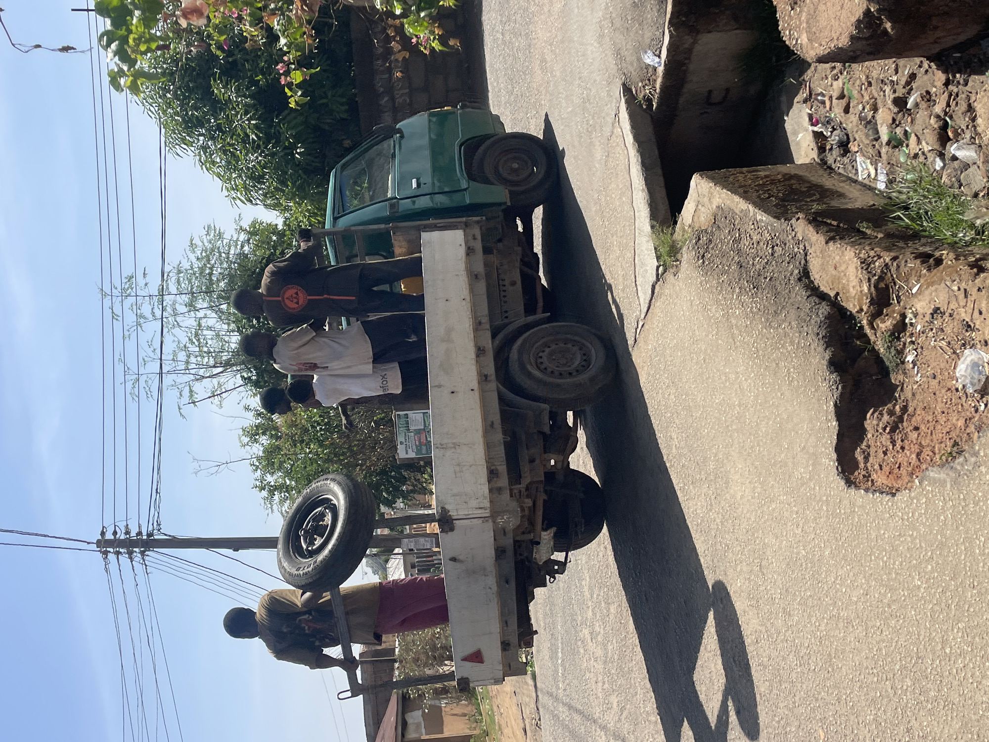 A bright and sunny street scene in Jos, Nigeria, depicts a green pickup truck with a modified, extended cargo bed and several men gathered around it. The truck is parked or slowly moving on a paved road, adjacent to a concrete drainage ditch.
In the truck's cargo area, at least four men are visible. One man wears a dark brown shirt featuring a circular emblem with text on its back. Another man in a white t-shirt has the word "Firefox" partially visible on his attire. A third man, dressed in a light brown shirt, is holding onto a metal railing at the rear of the truck, where a spare tire is also mounted. The men appear to be either traveling in the truck's open bed or in the process of loading/unloading.
The scene is under a clear blue sky, with bright sunlight casting prominent shadows from the truck and the men onto the road, suggesting it is midday or early afternoon. Lush green trees and foliage line the street. Overhead electrical wires are strung between utility poles in the background. The concrete drainage ditch shows signs of wear and contains some debris and dirt. The overall environment suggests a bustling, semi-urban setting.