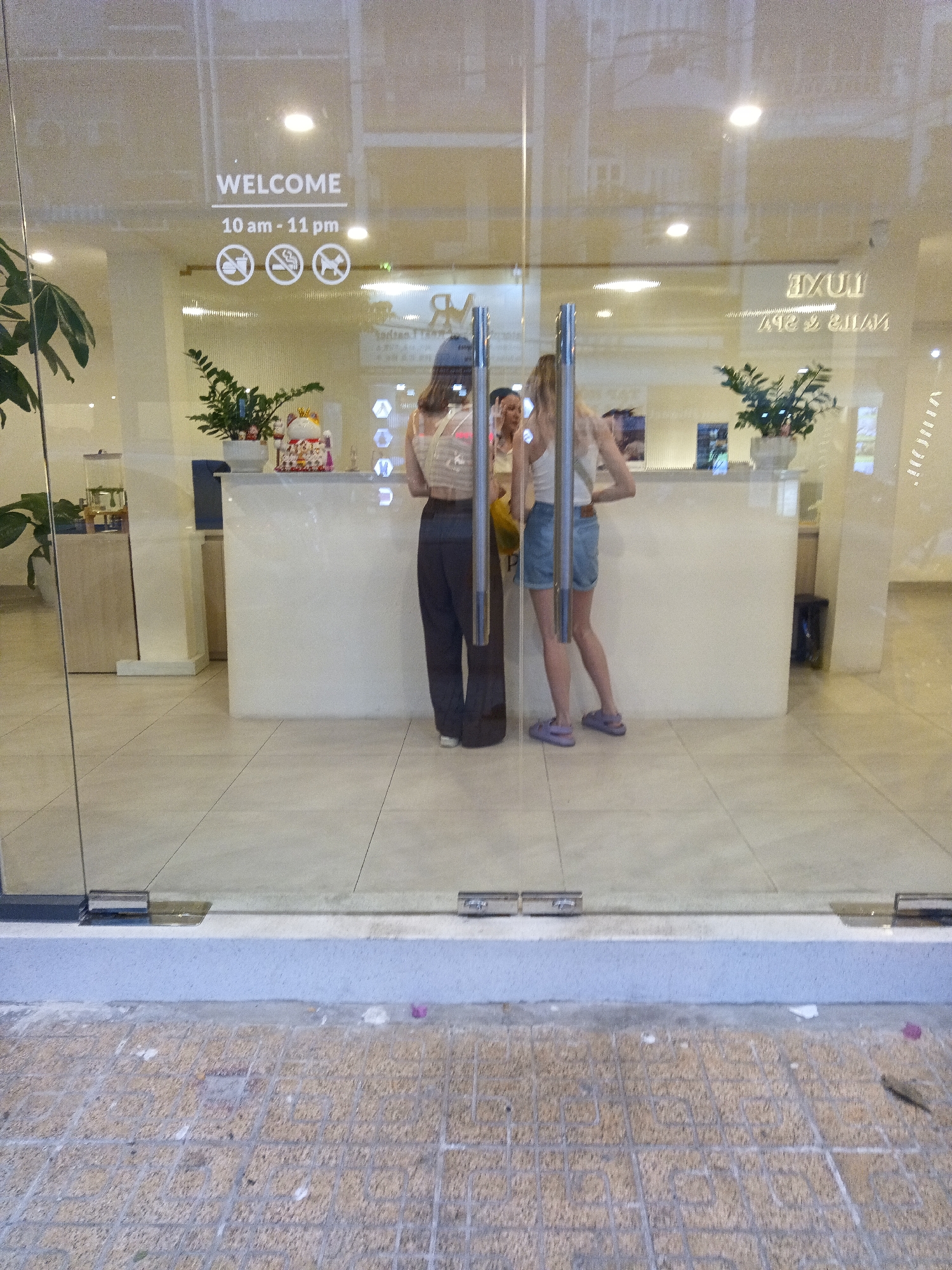The image shows the entrance to a business with a glass door and a large glass window. The sign on the window reads "WELCOME 10 am - 11 pm" along with three circular icons: a crossed-out cigarette, a crossed-out animal, and a crossed-out person. Inside the business, two women are visible at a reception desk. The woman on the left is wearing a hat and dark pants. The woman on the right is wearing shorts and a tank top. A potted plant and a decorative item are on the reception desk. The floor inside appears to be tiled. The exterior ground is paved with square tiles. The scene suggests a commercial establishment, possibly a salon or spa, operating in Nha Trang, Vietnam.