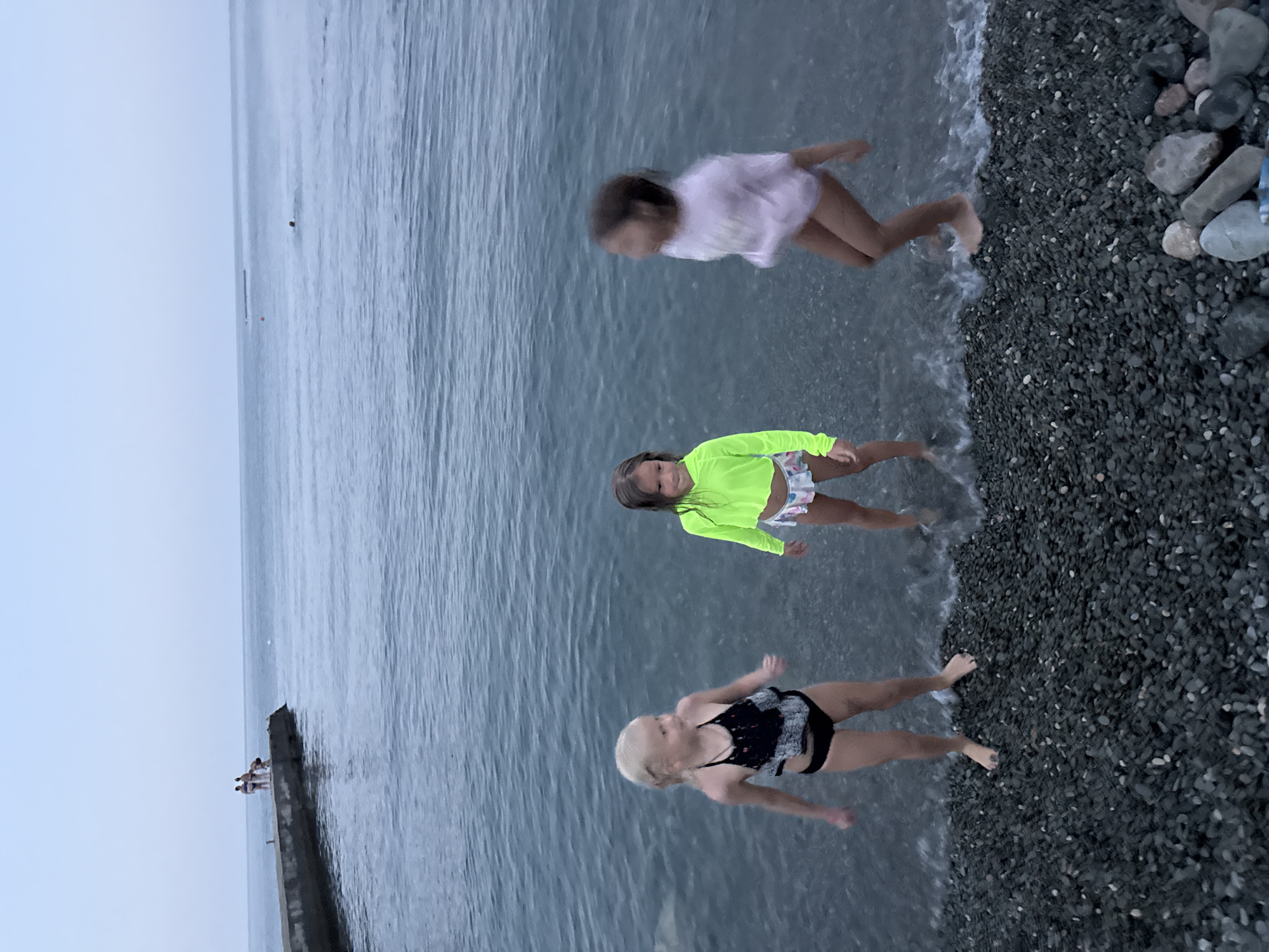 Three young girls are standing on a pebbled beach at the edge of the Black Sea in Partenit, Ukraine. The girl on the left, with blonde hair, is wearing a black and white swimsuit. The middle girl, with dark hair, is wearing a neon green long-sleeved top and patterned bottoms. The girl on the right, also with dark hair, is blurred due to motion and is wearing a pinkish top and light-colored bottoms. The water is calm with small waves lapping at the shore. In the background, a concrete pier extends into the sea, with a few figures standing on it. The sky is overcast, suggesting twilight or a cloudy day.