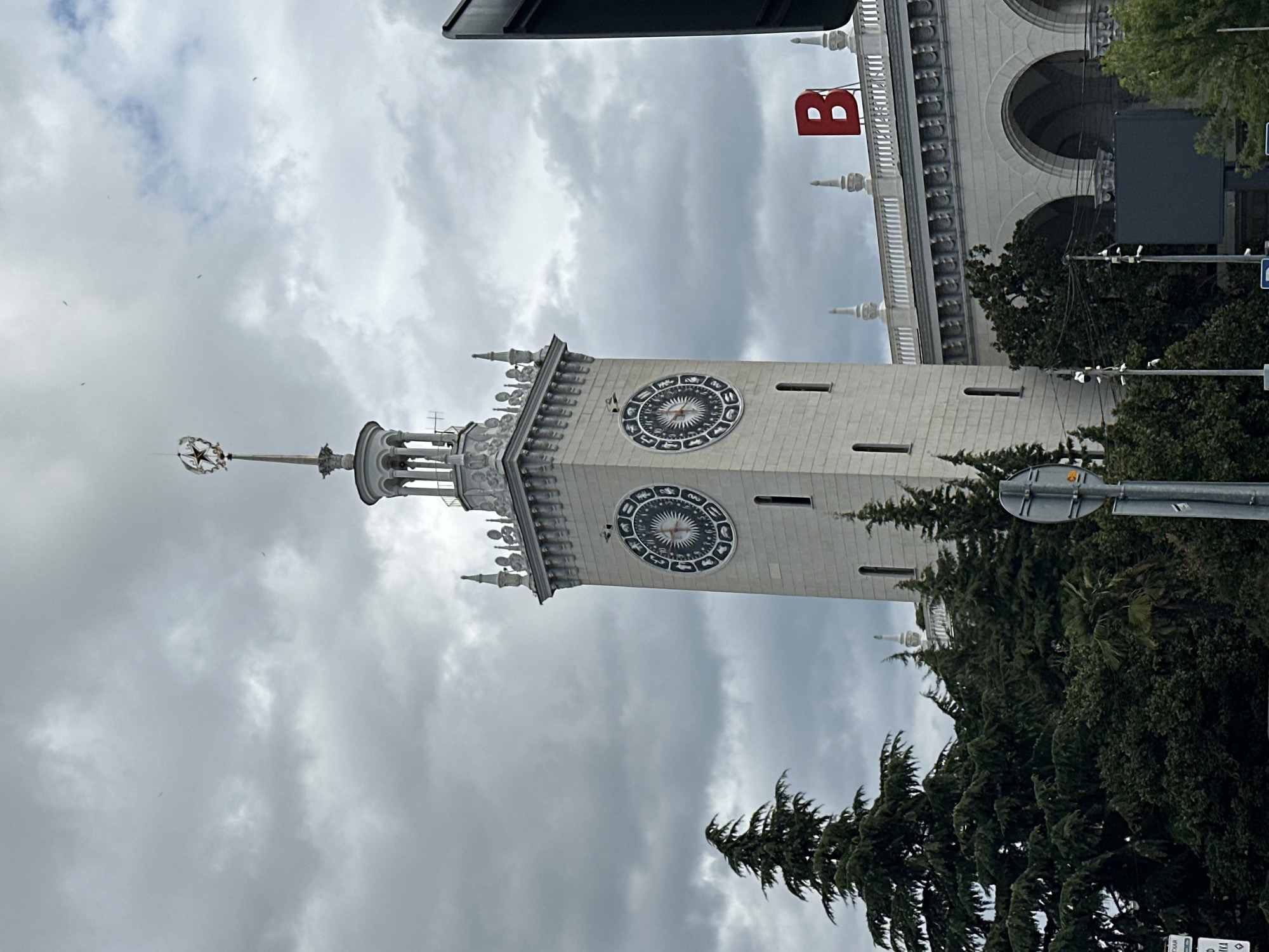 The image shows the Sochi Sea Port, a prominent landmark in Sochi, Russia. A tall, ornate clock tower with two large clock faces stands centrally in the frame, topped with a decorative spire. The tower is made of light-colored stone and features architectural details such as columns and decorative elements. Below the tower, a long, white building with arched entrances and a decorative balustrade is visible. A large red letter "B" is prominently displayed on the rooftop of this building. Lush green coniferous trees flank the left side of the tower, with more trees and foliage visible in the foreground and to the right, partially obscuring some parts of the scene. The sky is overcast with heavy, gray clouds, suggesting a cloudy or potentially rainy day. Several street signs and traffic lights are also present in the foreground. The scene appears to be an exterior shot of a public space, likely a transportation hub or a waterfront area.