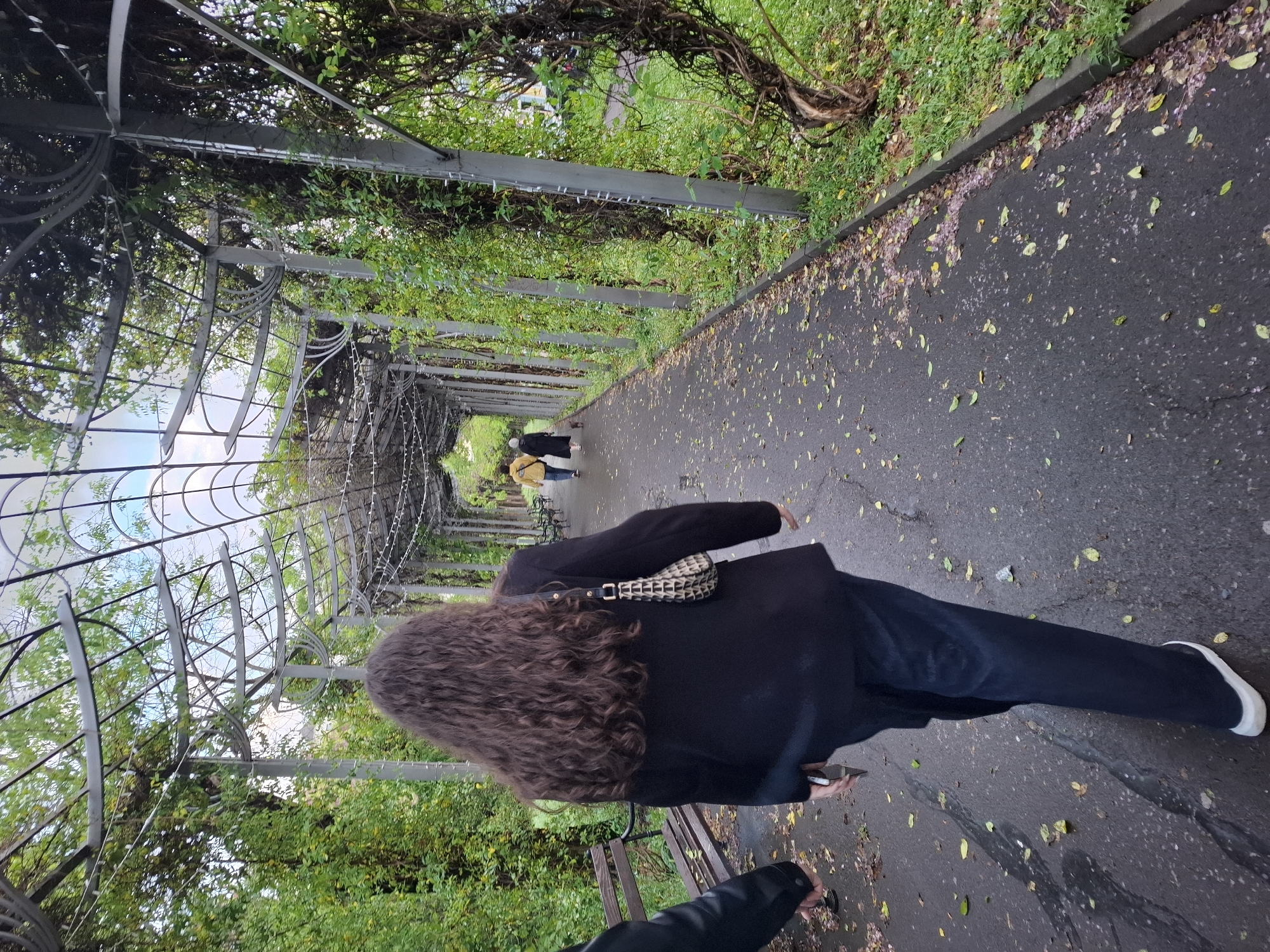 The photograph depicts an individual with long, curly brown hair walking along a pathway lined with an ornate metal pergola covered in greenery. The person is wearing dark clothing and a patterned bag. The path is paved and scattered with leaves. In the distance, two other individuals are walking in the same direction. The scene appears to be a park or garden setting. The location is most likely Burgas, Bulgaria, based on the known location of similar pergolas.
