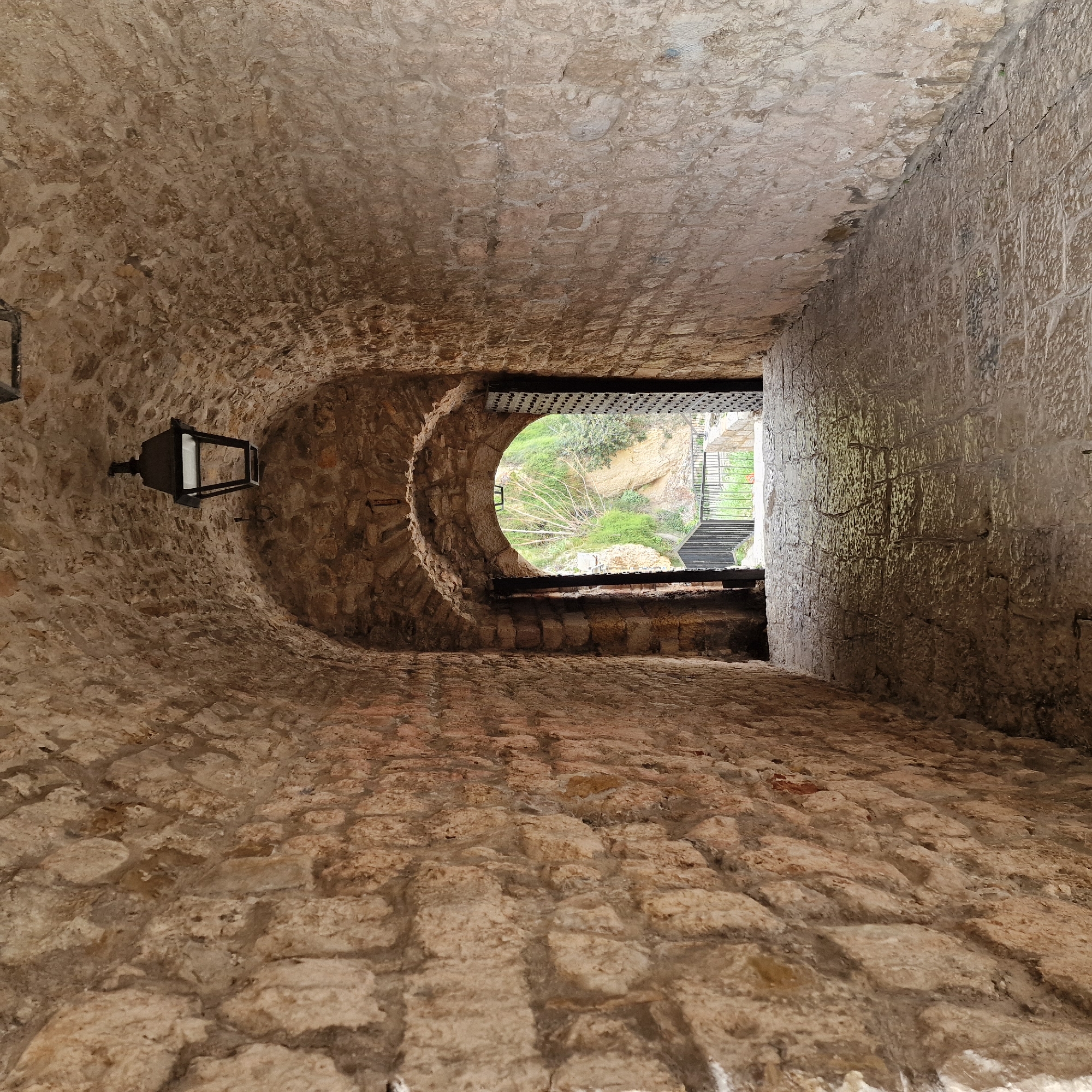 The image displays a long, arched stone tunnel, likely an entrance or passageway within the Old Town of Ulcinj - Ulqin, Montenegro. The tunnel's walls and vaulted ceiling are constructed from irregular, roughly hewn light-brown and reddish-brown stones, laid with visible mortar. The floor is paved with worn, rectangular stone slabs. A single, dark-colored, rectangular metal lantern is suspended from the center of the arched ceiling, approximately midway down the visible length of the tunnel. The lantern appears unlit.
The tunnel terminates in a large, arched opening, framed by two robust, dark-colored doors, heavily adorned with metal studs or panels, which are swung wide open and secured against the interior walls. Beyond this opening, an exterior scene is visible, illuminated by natural light. It features a steep, verdant hillside covered in dense green foliage, interspersed with lighter patches of exposed earth and rock. A prominent, light-barked tree with bare branches stands against the green backdrop. Immediately outside the archway, a black metal railing with vertical bars is visible, suggesting a protective barrier or a descending path. No individuals are present.