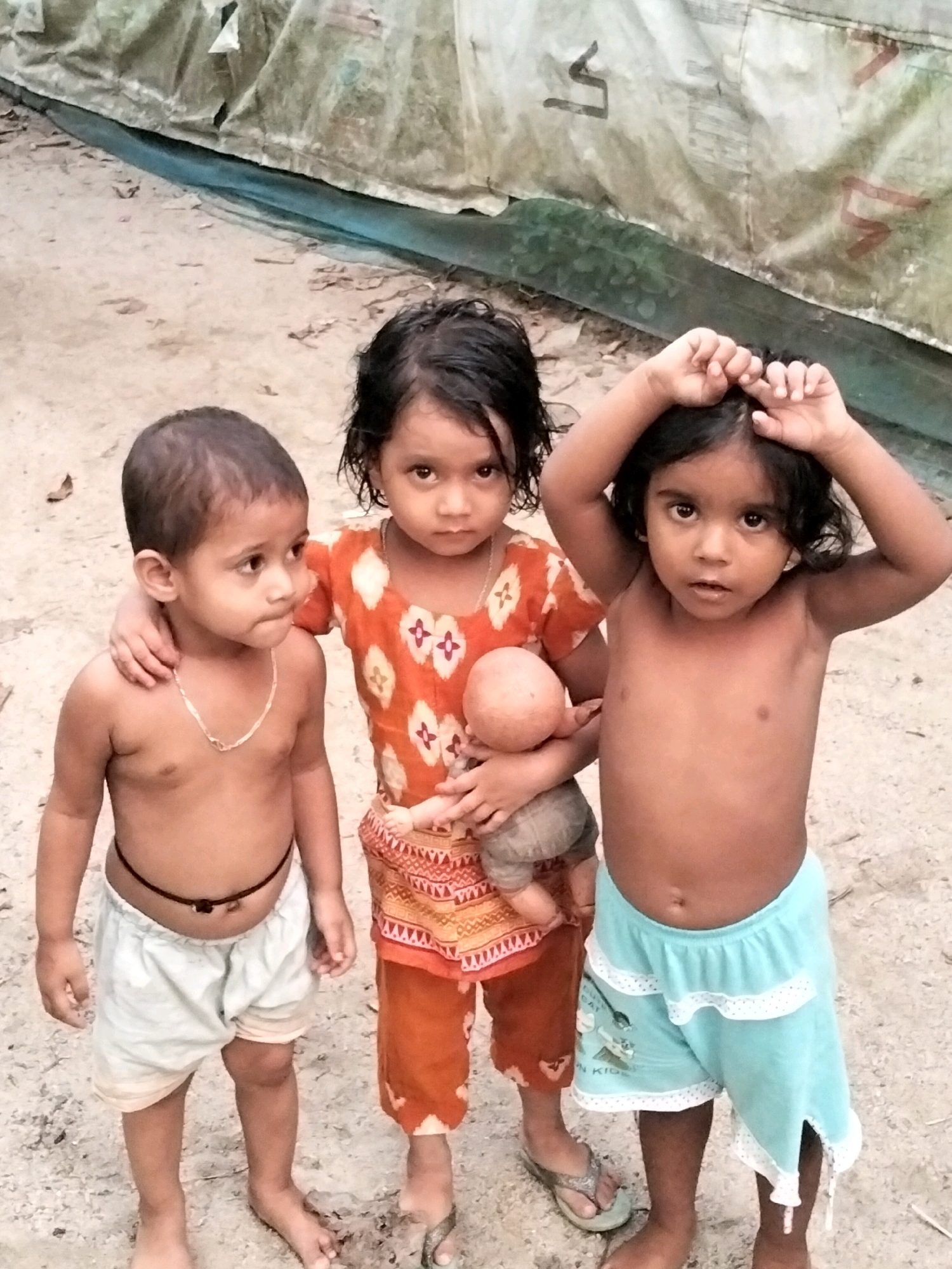 Three young children stand on sandy ground in front of a large, draped tarp. The child on the left is a boy with short hair, wearing light-colored shorts and a thin chain around his neck. He is looking to the side. In the center, a girl with dark hair is holding a doll and wearing an orange top with a patterned skirt. The child on the right is a girl with dark hair, wearing light blue shorts. She has her hands on her head and is looking at the camera. The tarp behind them appears to be part of a makeshift shelter, possibly in a rural area or a temporary settlement, consistent with observations from Bholagonj, Bangladesh. The ground is covered with sand and some debris.