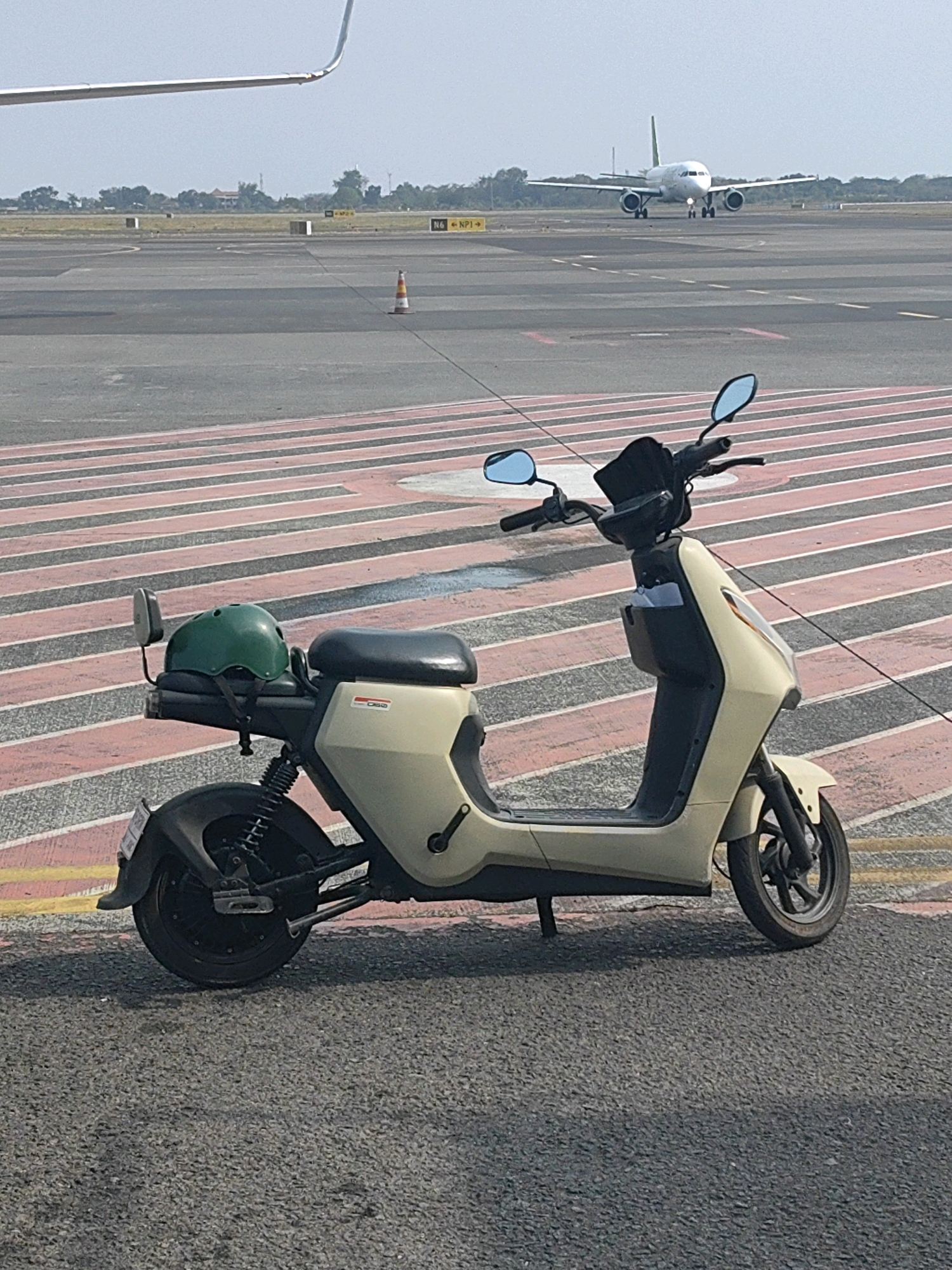 A light-colored electric scooter is parked on an asphalt surface next to a runway at an airport. The scooter has a green helmet secured to its rear rack. In the background, an airplane is taxiing on the runway. The scene appears to be located at an airport in Segoro Tambak, Indonesia, based on the visual cues of an airport environment and tropical vegetation. The sky is clear and the lighting suggests it is daytime.