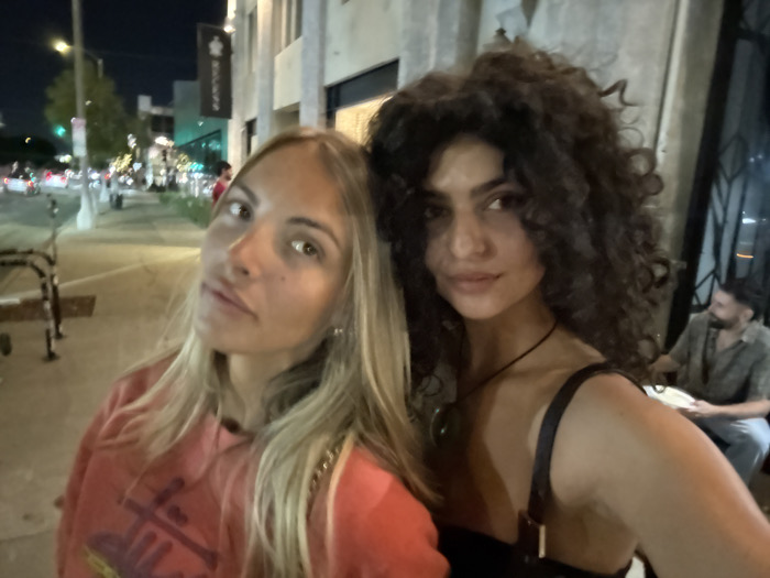 This image depicts an urban nighttime scene in an upscale commercial district of Los Angeles, United States of America, likely West Hollywood or Beverly Hills, near a Rolex boutique, indicated by a prominent black vertical banner with a white crown logo on a building facade.
In the foreground, two women pose for a photograph, seemingly a selfie, with the woman on the right holding the camera. The woman on the left has fair skin, long blonde hair, and light eyes; she wears a salmon-colored or coral hoodie with a dark graphic design on the chest and looks directly at the camera with a neutral expression. The woman on the right has darker skin, voluminous dark curly hair, and dark eyes; she wears a black top with a thick strap visible on her right shoulder and a dark necklace with a green pendant, smiling subtly at the camera.
In the mid-ground and background, an urban street environment is visible. To the left, a street light pole with a green traffic light is present, along with metal barriers on the sidewalk. Buildings with light-colored facades line the street, including the one featuring the aforementioned black banner with a white crown logo. Further down the street, additional streetlights illuminate the scene, and distant vehicle lights (red taillights and white headlights) suggest ongoing traffic. To the far right, partially obscured by the woman in the foreground, a bearded man with dark hair is seated at an outdoor table or counter, wearing a patterned short-sleeved shirt and blue jeans, looking towards the right side of the frame while holding a white object. The pavement is visible in the immediate foreground, and the sky is dark, indicating night.