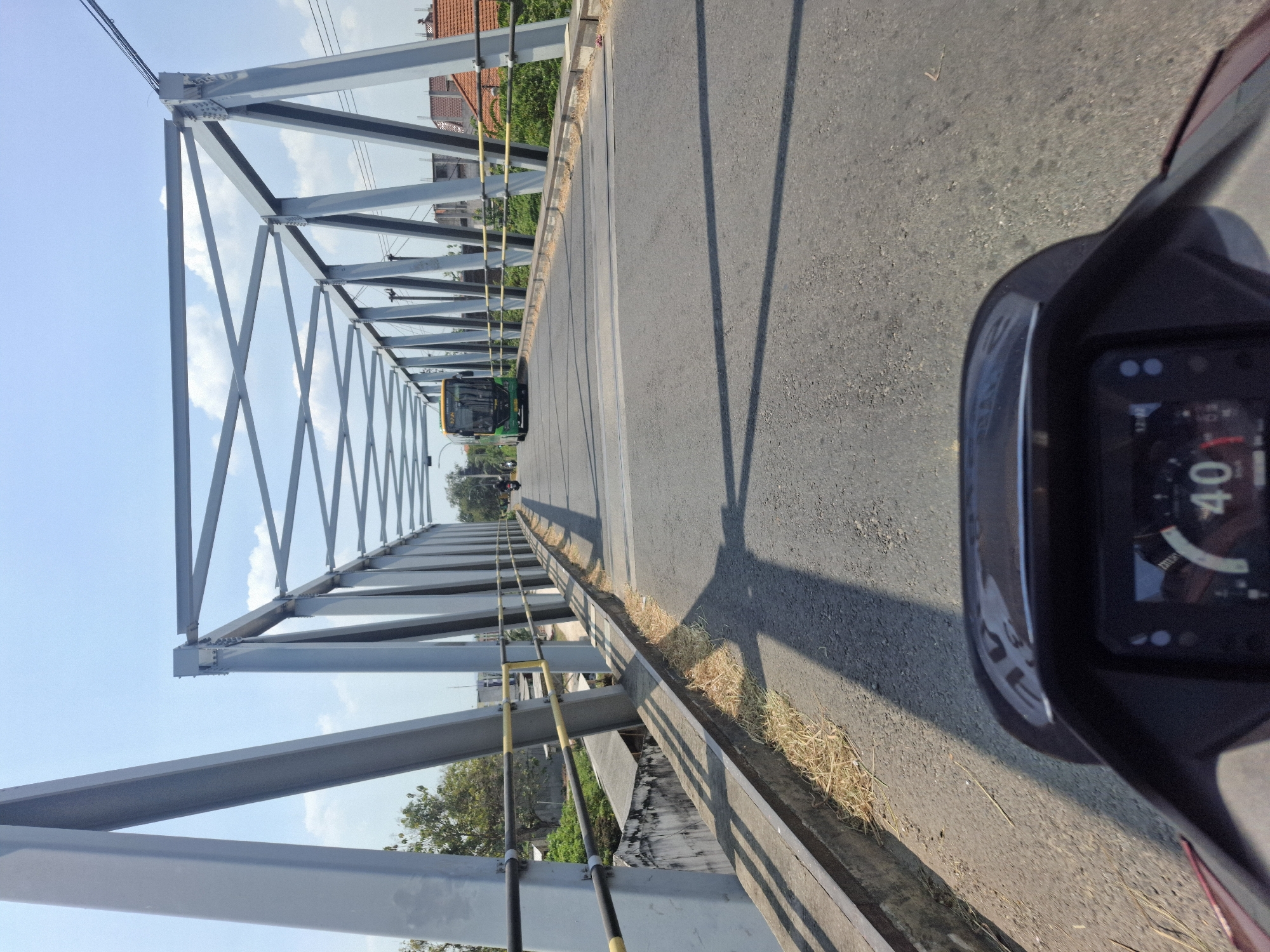 The image captures a first-person perspective from a motorcycle traveling across a bridge in Karangcangkring, Indonesia. The bridge has a prominent grey metal truss structure overhead, with diagonal supports and horizontal beams forming a geometric pattern against a clear blue sky. The roadway is asphalt and appears to be in good condition, with visible lane markings.
On the bridge, a green and yellow bus is seen in the distance, traveling in the same direction as the motorcycle. To the left of the road, there is a concrete retaining wall with dry grass growing along its edge. The surrounding environment shows vegetation, buildings with red tile roofs, and electrical wires.
The motorcycle's dashboard is partially visible in the foreground, displaying a speed of 40. The overall scene depicts a sunny day, suggesting a typical transportation route within the Indonesian city.