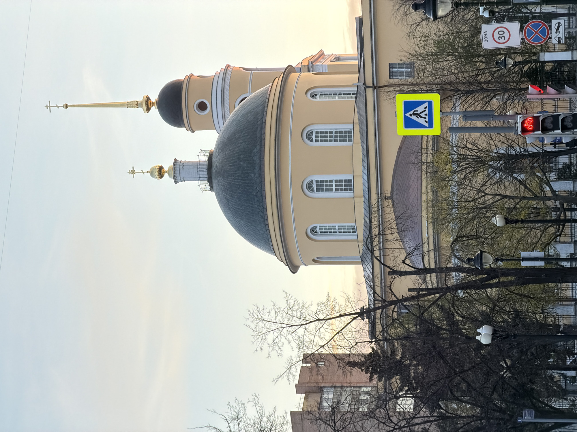 The image shows a section of a building with two domes, likely an Orthodox church. The building is light yellow with white window frames. The domes are dark gray, with a gold-colored spire topping one.
Foreground elements include trees with bare branches, a yellow and blue pedestrian crossing sign, a street lamp, and a traffic light showing a red light. Other street signs, including a speed limit sign in Russian, are visible in the background. There are other buildings behind the trees and church.
The image is likely taken in Moscow, Russia, given the architectural style of the church and the Russian-language signs.