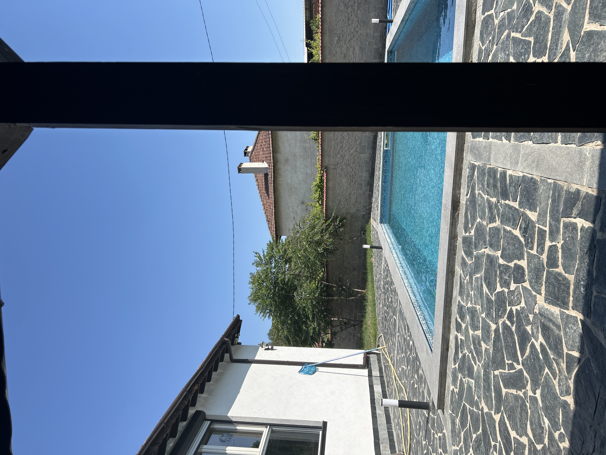 The image displays an outdoor scene on a clear, bright sunny day, viewed from beneath a dark horizontal beam, likely part of a pergola or overhead structure. The foreground features a spacious patio paved with dark, irregularly shaped flagstones, bordering a rectangular swimming pool with bright blue mosaic tiles. A second, similar swimming pool is visible further back, separated by a dividing wall.
To the left, a white stucco house with a distinct brown, scalloped tiled roof is prominent, featuring a window and a rain gutter system. A blue pool cleaning net hangs on the house's wall, and a yellow garden hose is coiled on the ground next to an outdoor light fixture. Further in the background, another house with a light-colored wall and a reddish-brown tiled roof, adorned with two chimneys, is partially obscured by a large, lush green tree. Utility lines are faintly visible stretched across the expansive blue sky.
The scene suggests a warm, peaceful atmosphere, indicative of midday or early afternoon due to the strong sunlight and crisp shadows. No individuals are present, and no text is visible within the frame. The overall setting is a well-maintained residential property with recreational facilities.