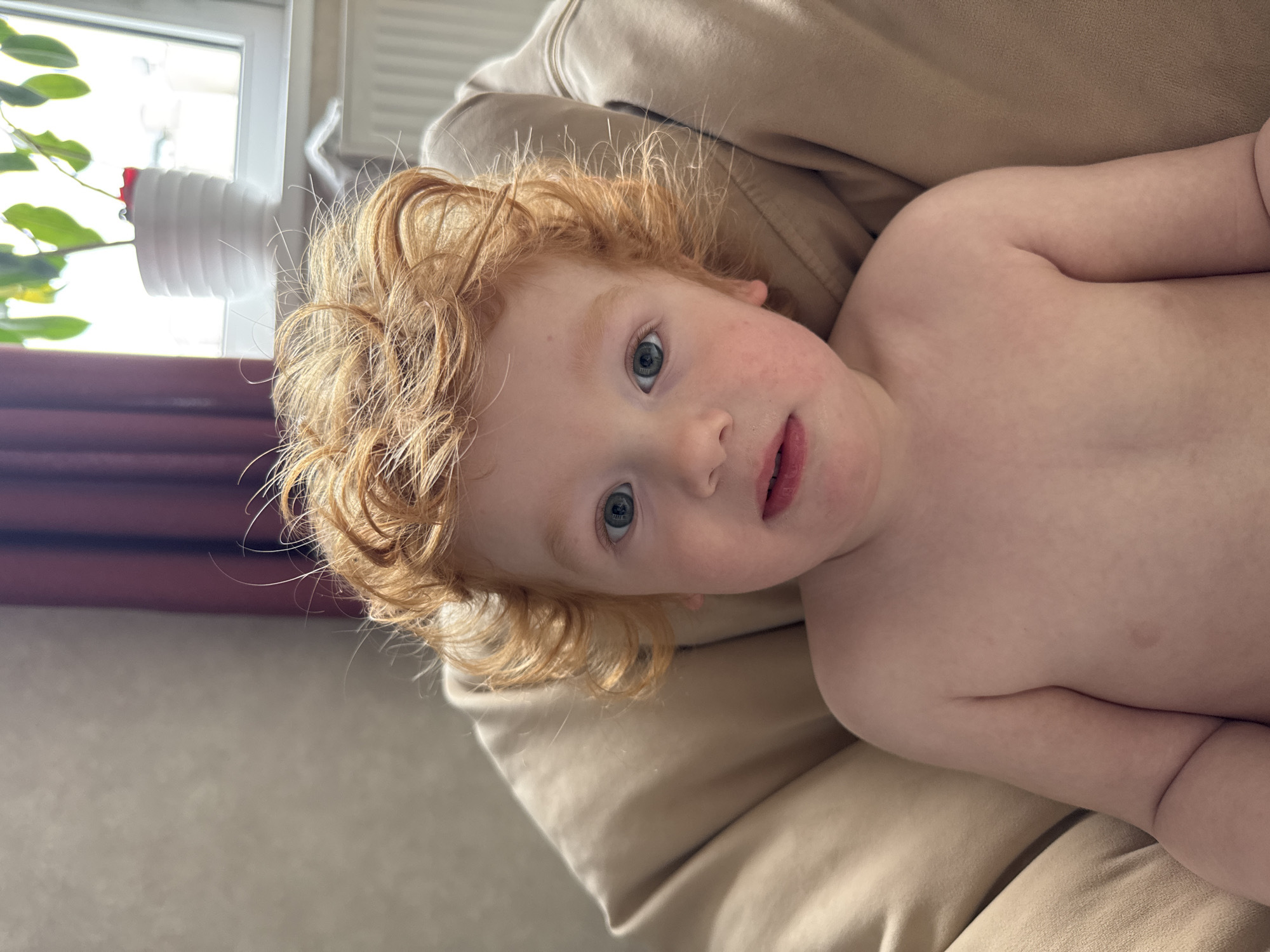 A close-up, slightly rotated image shows a fair-skinned child with red, curly hair lying on a light brown couch or cushion. The child has light eyes and is looking at the camera. The child is unclothed from the chest up. A plant in a white pot is visible in the background, near a window with blinds. The location is inside a building. There is no indication of being in the city of Saint Petersburg, Russia.