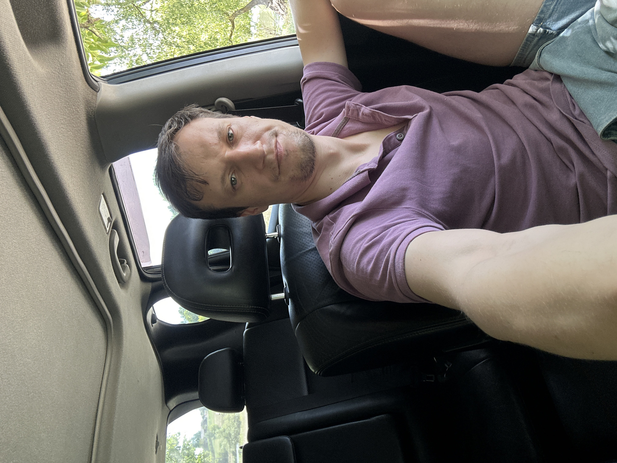 In the city of Makiv, Ukraine, a man is pictured inside a vehicle. He is seated in the front passenger seat, his right arm resting on the car's interior. He is wearing a purple polo shirt and denim shorts. Through the car window, lush green trees and foliage are visible, suggesting an outdoor, possibly rural setting. The car's interior, including black leather seats and a sunroof, is also discernible. The man is looking directly at the camera, his expression neutral.