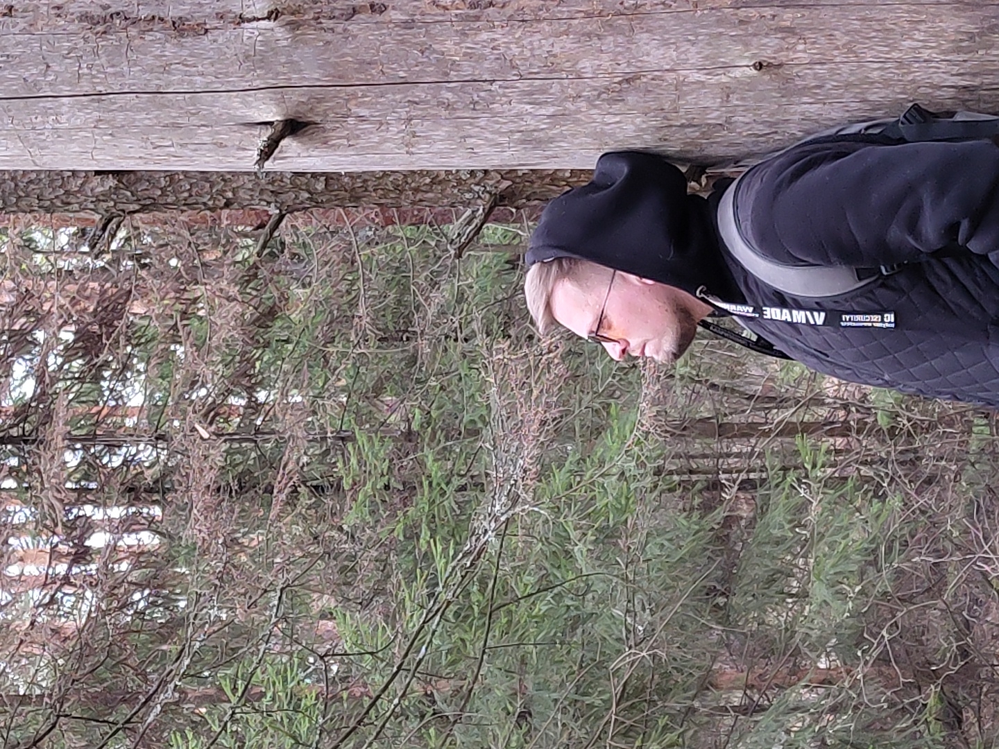 A fair-skinned person with blonde hair, wearing a black hooded jacket, glasses, and a backpack, stands against a backdrop of dense greenery and a wooden structure. The location is in Прикамский, Russia. The environment features both dense plant life and artificial structures.