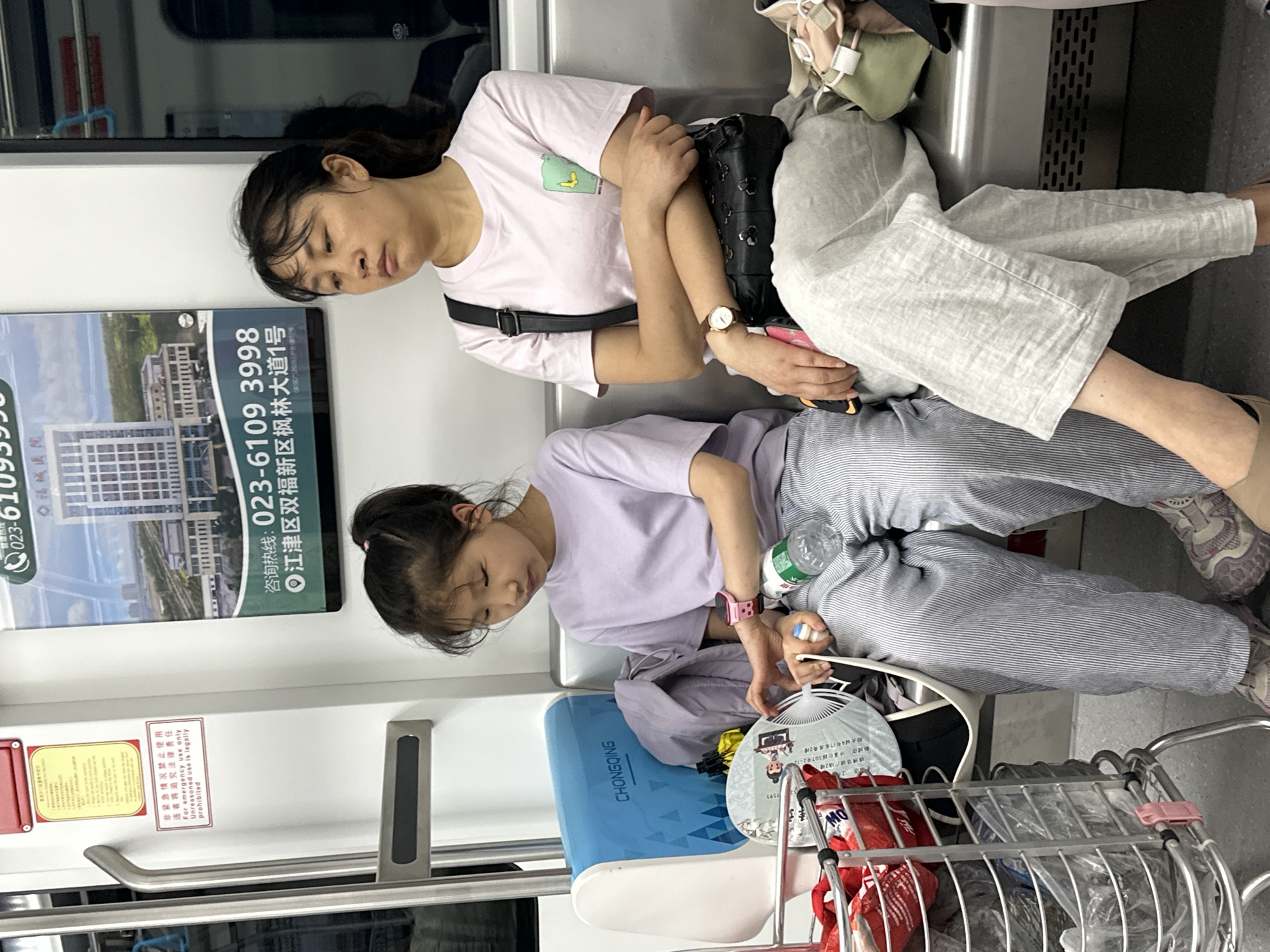 The image shows an adult woman and a young girl sleeping on a public transit seat, likely a train, within the city of Jiulongpo District, China. The adult is leaning against the seat's back, wearing a light pink shirt and light grey pants, with a black bag on her lap. The young girl is seated next to her, wearing a light purple shirt and striped grey pants. A water bottle rests near her hands, and a shopping cart with several items including a fan is placed adjacent to them. An advertisement is visible on the train car wall, featuring the contact information for a hospital in Jiangjin District.