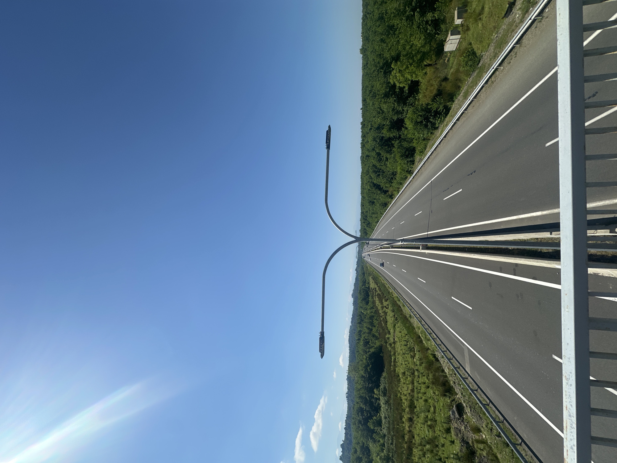 The image depicts a highway in Ureki, Georgia, under a clear blue sky with a bright sun casting lens flare. The highway, divided by a median, features multiple lanes marked with white lines and stretches into the distance, bordered by lush green vegetation and dense trees on either side. Two large, modern streetlights arch over the highway. In the foreground, a metal railing suggests the viewpoint is from an overpass or pedestrian bridge. A single white car is visible on the distant highway. The scene is tranquil and devoid of human presence beyond the implied observer.