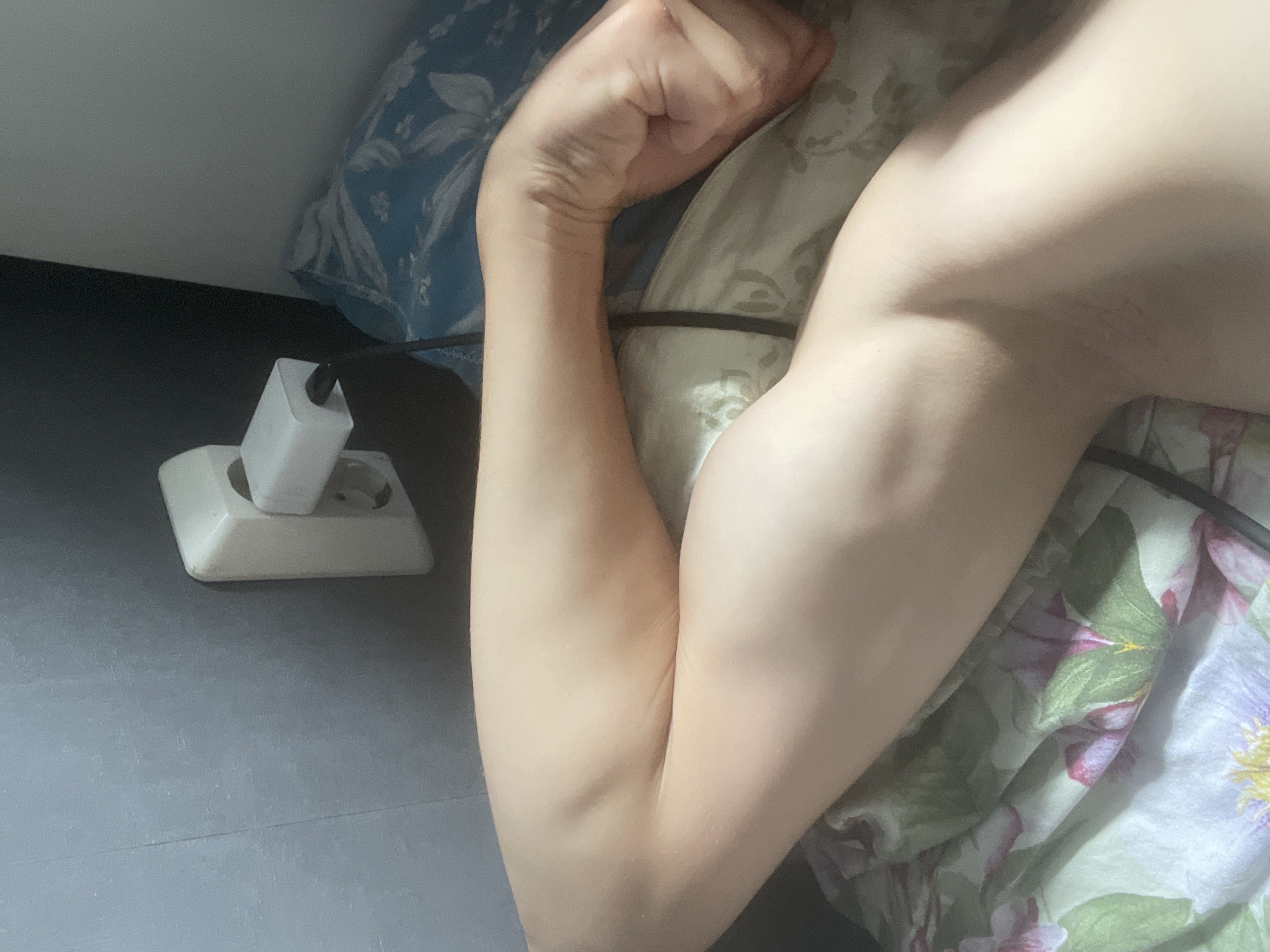 A close-up shot focuses on a person's arm, bent at the elbow, resting on floral-patterned bedding. The arm displays well-defined muscles, suggesting a toned physique. In the background, a white electrical outlet with a charger is visible on a gray wall. The lighting is soft, casting subtle shadows and highlighting the texture of the skin. The scene appears to be indoors, possibly in a bedroom, with the overall impression being one of relaxed repose.