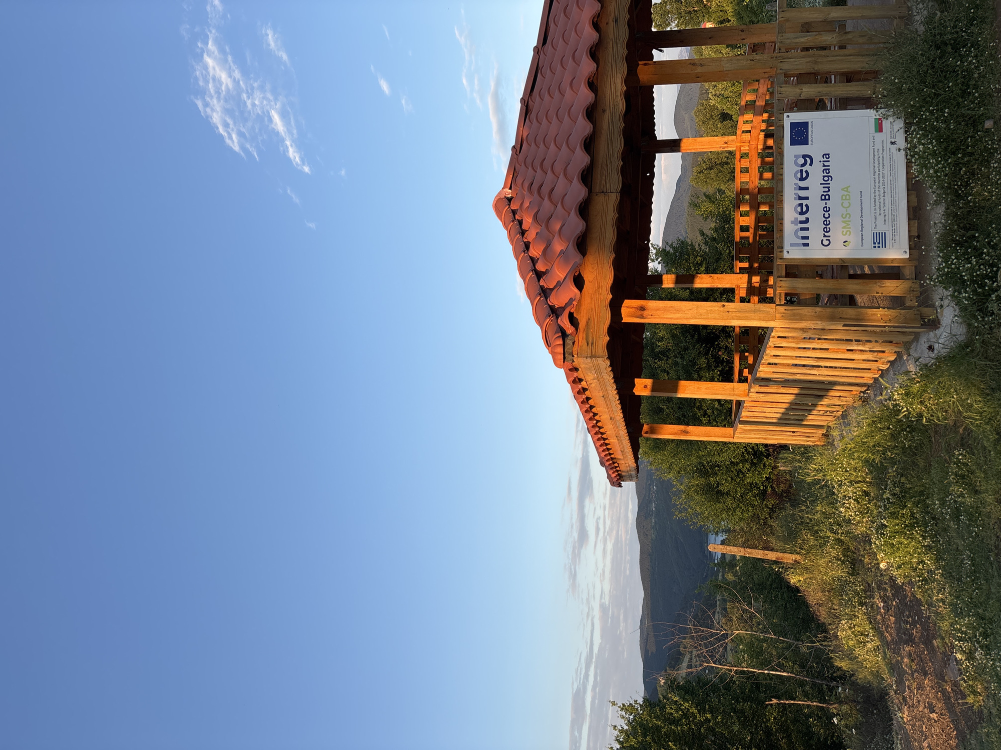 A wooden gazebo with a red-tiled roof stands on a grassy hill overlooking a valley in Ribartsi, Bulgaria. The gazebo is illuminated by the warm glow of the setting sun, casting long shadows across the ground. A sign inside the gazebo indicates it is part of the "Interreg Greece-Bulgaria SMS-CBA" project, funded by the European Regional Development Fund. The surrounding environment is lush with green trees and wildflowers. The sky is a clear blue with a few wispy clouds. The overall scene evokes a sense of peace and natural beauty.