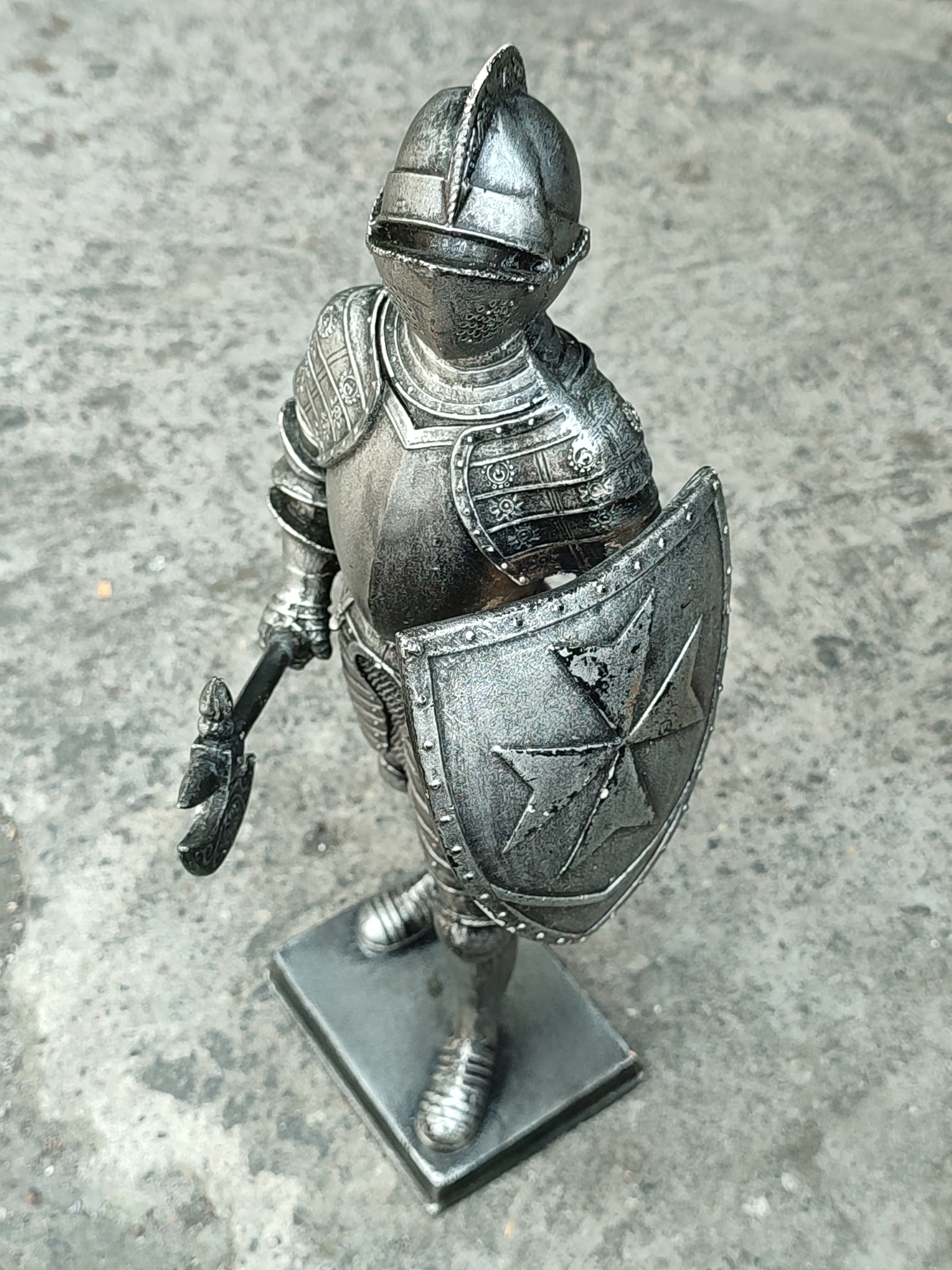 A detailed, metallic figurine of a knight stands on a grey, textured surface, likely concrete. The knight is depicted in full armor, including a helmet with a visor, a breastplate, pauldrons, and gauntlets. The armor is intricately detailed with patterns and ridges. The knight holds a shield on its left side, emblazoned with a white eight-pointed cross on a dark background, characteristic of the Maltese cross. In its right hand, the knight holds a weapon that appears to be a battle axe. The figurine is mounted on a rectangular base. The background is out of focus, showing a mottled grey texture consistent with an outdoor urban environment. The lighting suggests daylight. There are no people or any indication of the city of Petiken, Indonesia.