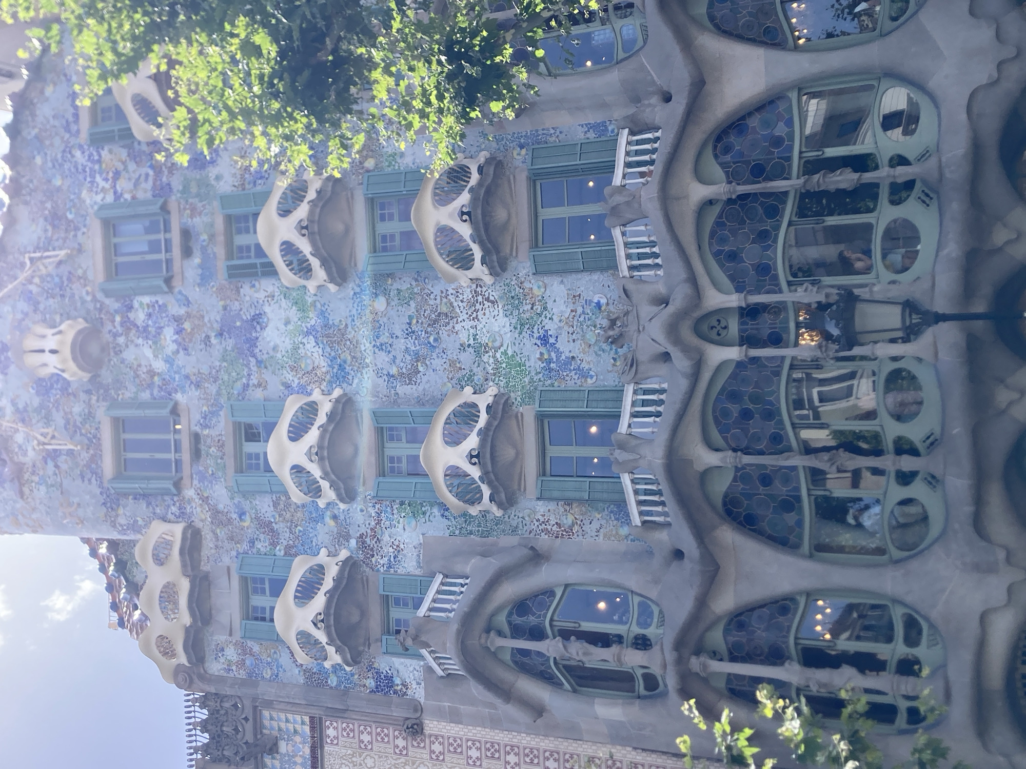 The image depicts the exterior of Casa Batlló, a building in Barcelona, Spain, designed by Antoni Gaudí. The facade is a vibrant mosaic of colorful tiles and sculpted stone, creating an organic, flowing aesthetic. Several unique balconies, resembling bones or masks, protrude from the building. The windows are also distinct, with rounded frames and intricate stained glass. Lush green foliage from surrounding trees partially obscures the upper portions of the building, suggesting it is a sunny day and possibly spring or summer. The light suggests it is daytime, likely morning or afternoon. The scene captures the architectural artistry and playful design of Gaudí's work. There is no visible text in the image.