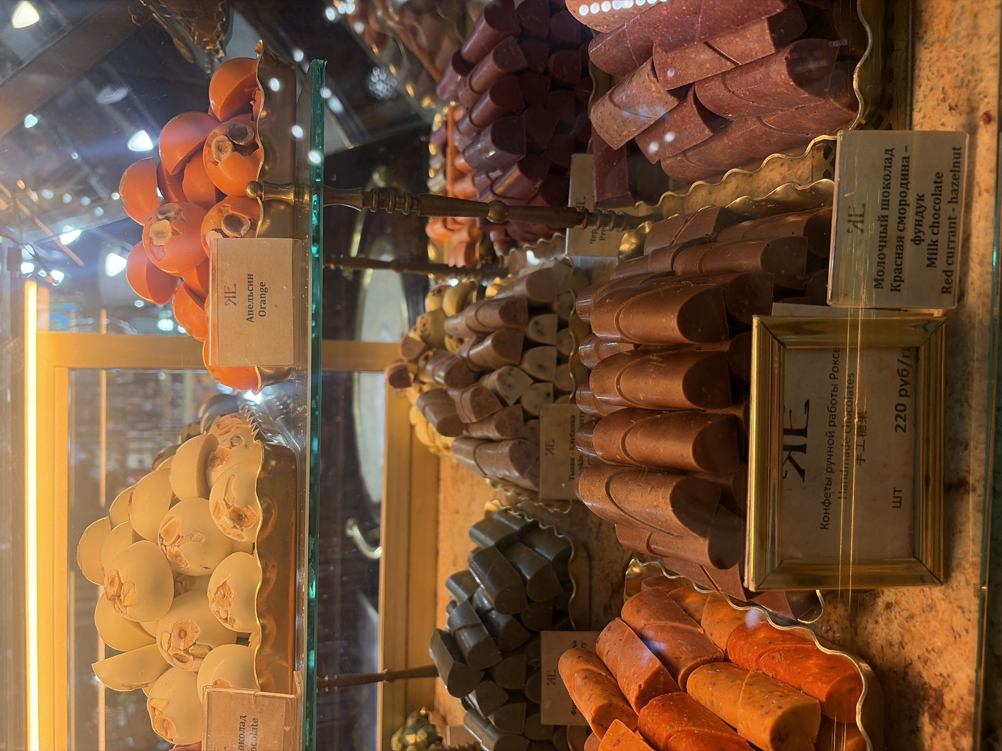 The image is a close-up, eye-level shot of a display case filled with various types of chocolates. The chocolates are arranged on golden trays and shelves, illuminated by warm, artificial lighting that casts a glow on the confectionery.
In the upper left, a tray holds white chocolates with visible hazelnut pieces embedded in them. A small sign next to them reads "шоколад" and "chocolate". To the right of these, another tray is filled with orange-colored chocolates, some of which are halved, revealing a hazelnut inside. A sign with Cyrillic text and the English word "Orange" is placed in front of this tray.
Below these, on the left, a tray contains dark green, rectangular chocolates. Next to them, a tray is filled with lighter brown, ridged chocolates. A sign with a logo and Cyrillic text is in front of the green chocolates, and another sign with similar branding is in front of the ridged chocolates, which appear to have lighter speckles within them.
In the foreground, a tray on the left is piled high with orange-colored, cylindrical chocolates. To the right of these, a tray holds a large quantity of dark brown, cylindrical chocolates, some of which are cut in half. Behind these, a tray is filled with dark red, rod-shaped chocolates.
A framed sign in the lower center of the image displays a logo and text in Cyrillic and English, stating "Конфеты ручной работы Рокс", "Handmade chocolates", and Chinese characters, followed by "шт 220 руб/г". To the right of this, another sign with the same logo and Cyrillic text is accompanied by the English text "Milk chocolate Red currant - hazelnut".
The overall environment appears to be a high-end confectionery shop or a chocolate boutique. The display case has glass panels, and reflections of the surrounding environment are visible, including wooden structures and other display elements. The polished surfaces and the rich colors of the chocolates suggest a luxurious and tempting presentation. The presence of Cyrillic text indicates the location is likely in Russia, and the specific branding and context suggest it could be a shop in Saint Petersburg, Russia, known for its patisseries and confectioneries.