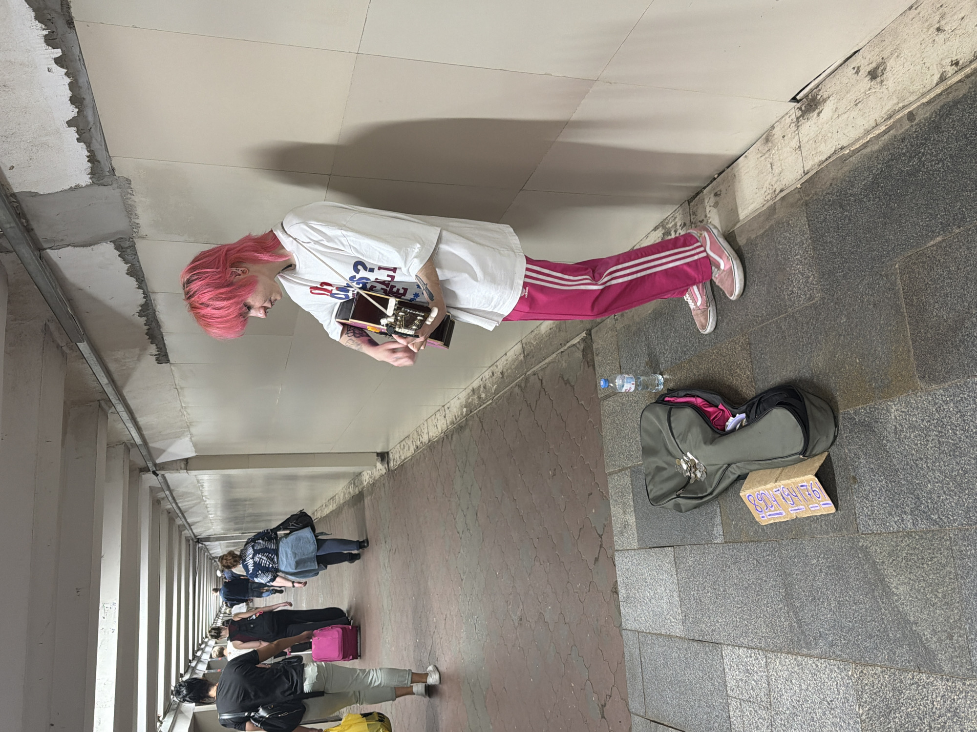 An individual with bright pink, mullet-style hair is playing an acoustic guitar in a subway passageway. The person is wearing a white t-shirt with a graphic print and vibrant pink sweatpants with white stripes. They are standing and leaning against a cream-colored wall. In front of them, on the ground, is a gray guitar case with some coins and a folded piece of cardboard with writing on it. A water bottle is also visible next to the guitar case. The floor of the passageway is made of reddish-brown patterned tiles, transitioning to gray granite-like tiles. In the background, several people are walking in the opposite direction, some carrying luggage, indicating a public thoroughfare. The environment appears to be an indoor, utilitarian passageway, possibly a pedestrian underpass or a section of a transit station. The lighting is artificial and somewhat dim. No specific markings clearly identify the location as Nizhny Novgorod, Russia.