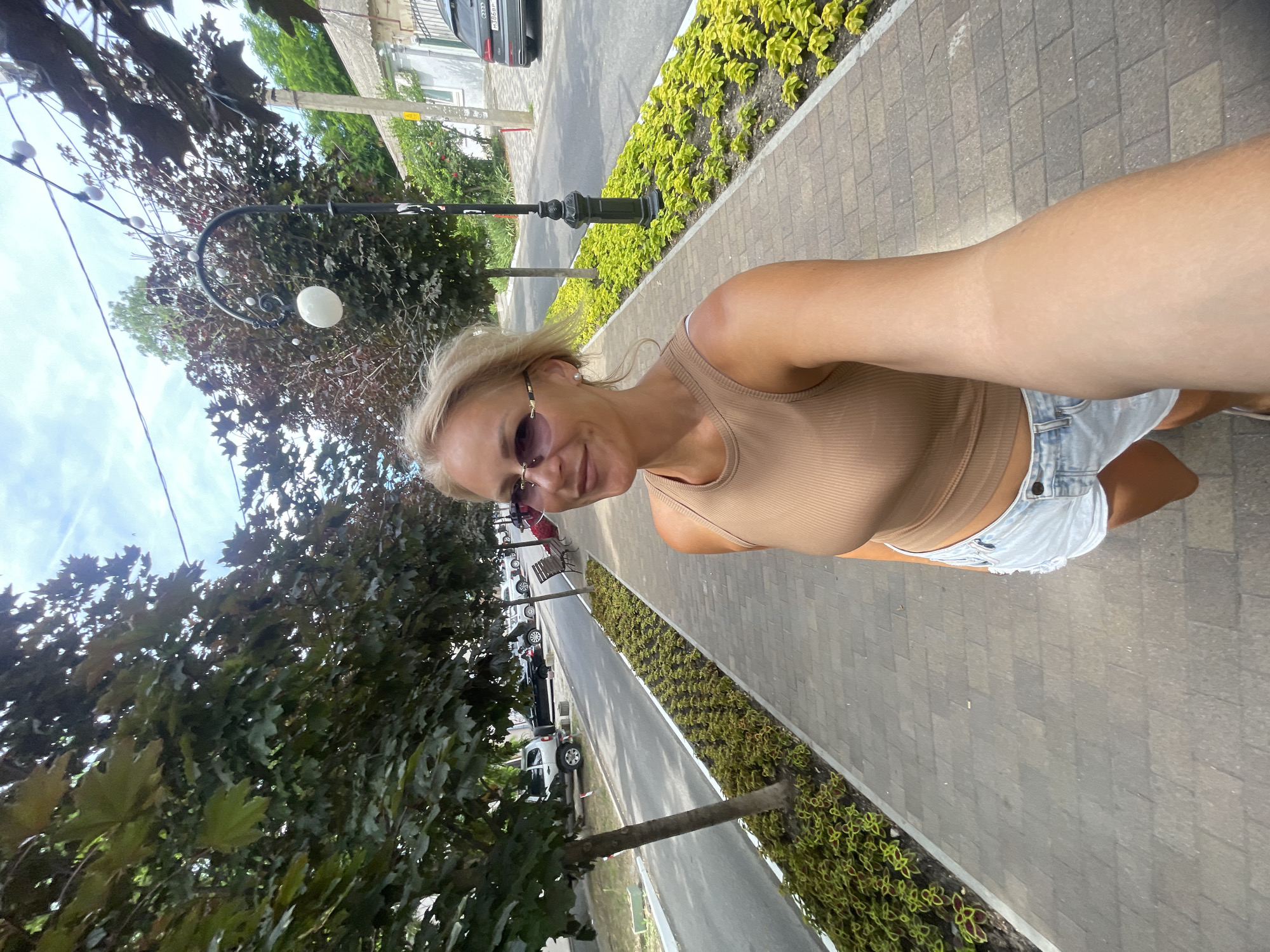 The original image shows a blonde woman with sunglasses taking a selfie while walking on a brick-paved sidewalk. She is wearing a tan tank top and denim shorts. The sidewalk is lined with trees and a flowerbed. A street with parked cars is visible to the left, and houses and a street lamp are on the right. This scene is consistent with a promenade or street in Anapa, Russia, a coastal city known for its pedestrian-friendly areas.