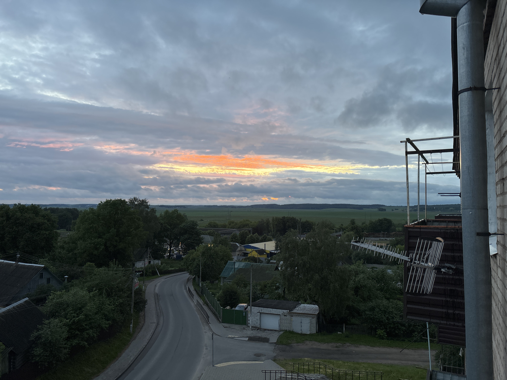 The image captures a view of a small town, likely in or near Smalyavichy, Belarus, at dusk. The sky is filled with dramatic clouds, with streaks of orange and pink light indicating the setting or rising sun breaking through. In the foreground, the right side of the frame shows a brick building with a large metal downspout and an attached antenna structure. Below, a winding asphalt road curves through a residential area characterized by scattered houses with dark roofs and surrounding trees. Some of the houses appear to be simple, rural dwellings. Beyond the houses, a wide expanse of green fields stretches to the horizon, under the cloudy sky. The overall impression is one of a quiet, rural evening scene.