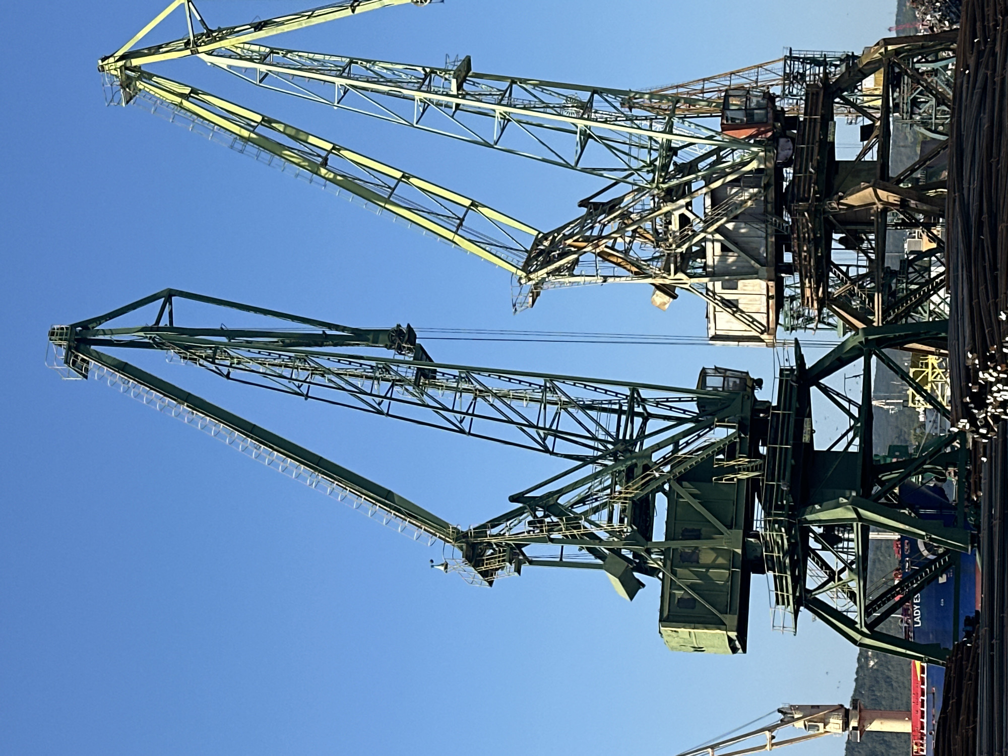 Two large, industrial harbor cranes dominate the frame against a clear blue sky. The crane on the left is painted a dark green, while the one on the right has a lighter green and yellow color scheme. Both cranes are tall, metal structures with complex lattice frameworks and extended jibs. The crane on the left has a rectangular control cabin attached to its tower. The crane on the right also has cabins and a more weathered appearance. In the foreground, a large pile of dark, metallic rods or pipes is visible, suggesting a port or industrial storage area. Behind the cranes and to the left, a blue ship with the name "LADY ES" visible on its hull is docked. In the background, a distant, hazy shoreline with hills can be seen. Another smaller, lighter-colored crane is partially visible on the far left of the image. The scene appears to be a sunny day in an industrial port environment.