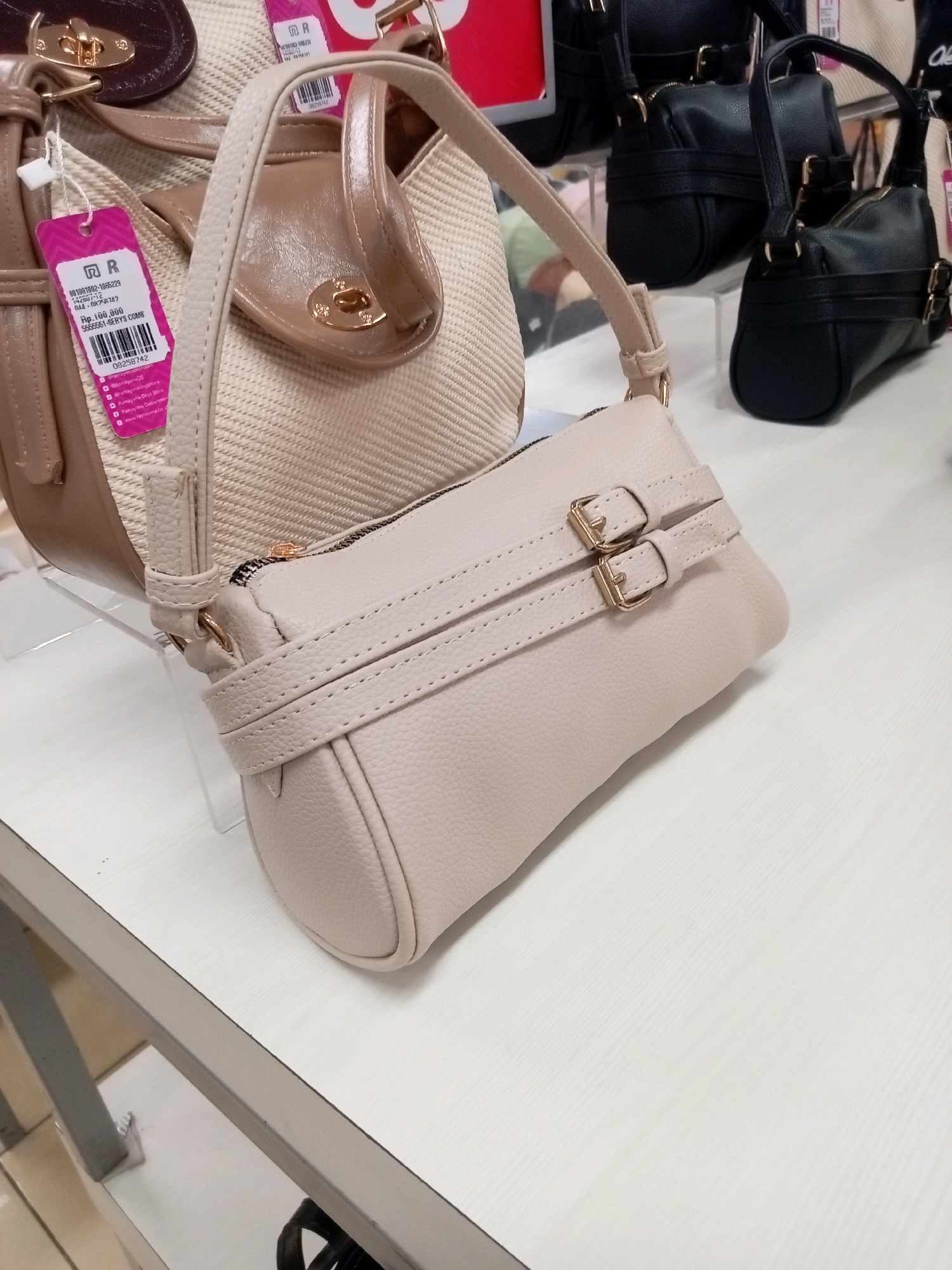In Batam, Indonesia, a retail display features several handbags. The primary subject is a pale pink, cylindrical handbag with a textured leather-like finish. It has two parallel straps across its front, each secured with a gold buckle. A gold zipper runs along the top. Behind this handbag, a larger, light beige woven tote bag is visible, adorned with beige faux leather accents and a gold clasp. To the right, two black, structured handbags with similar gold buckle detailing are displayed. A pink price tag with a barcode is attached to the woven tote bag. The handbags are arranged on a light-colored, textured surface, possibly a shelf or countertop.