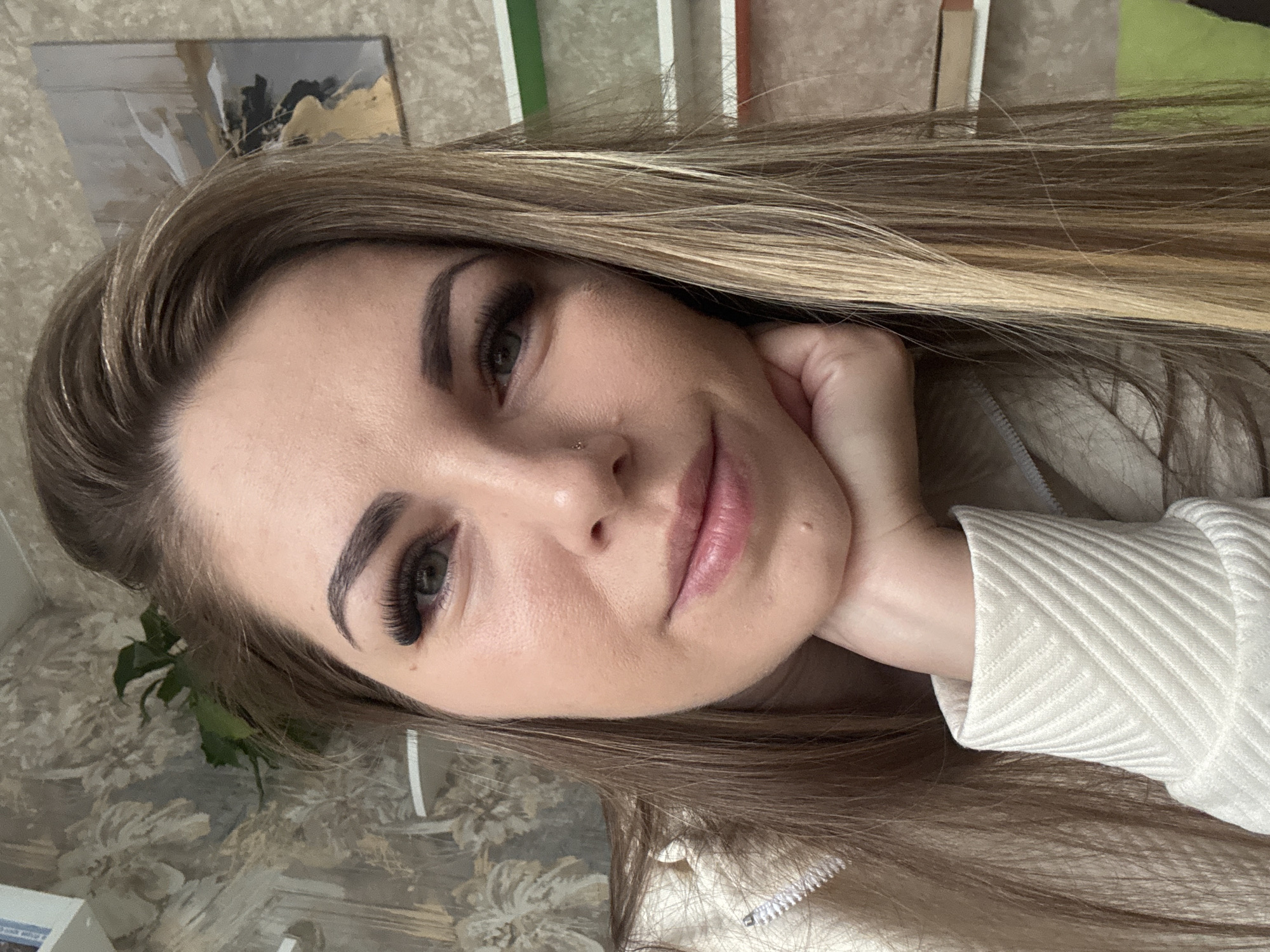 A woman with blonde hair and extended eyelashes rests her head on her hand, looking directly at the camera. She has a nose piercing. She wears a light-colored, textured garment. The background includes patterned wallpaper and a painting. The location is Раздолье, Russia.