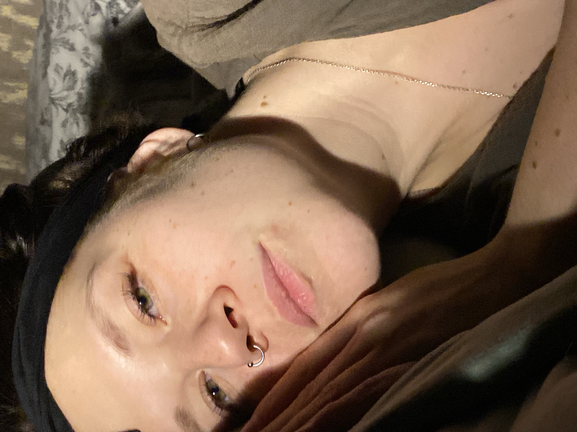 The image shows a person with fair skin, hazel eyes, and brown hair wearing a black headband. The person has a nose ring and a delicate silver chain around their neck. They are lying down, partially covered by a brown fabric, possibly a blanket or clothing. The background is dark, with a hint of floral patterns suggesting bedding or curtains. There is no indication that the image was taken in Chaikovsky, Russia.