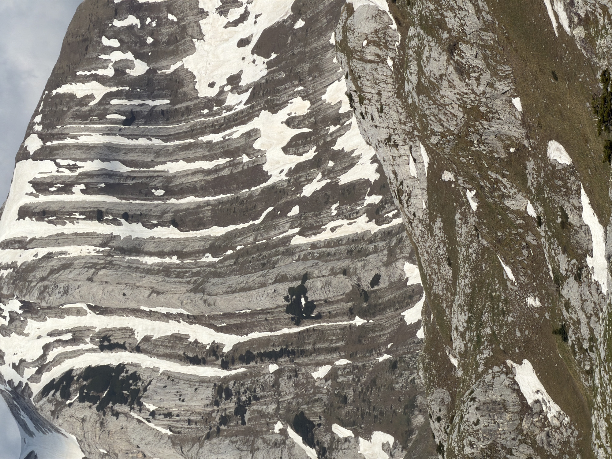 The image depicts a mountain landscape in Boričje, Montenegro. The left side of the image features a layered rock formation, with bands of gray stone interspersed with patches of white snow. The right side of the image shows a more rugged, uneven terrain, with a mix of gray rocks, green vegetation, and scattered patches of snow. There are no people visible in the scene. The environment appears to be a high-altitude, rocky area, typical of mountainous regions.