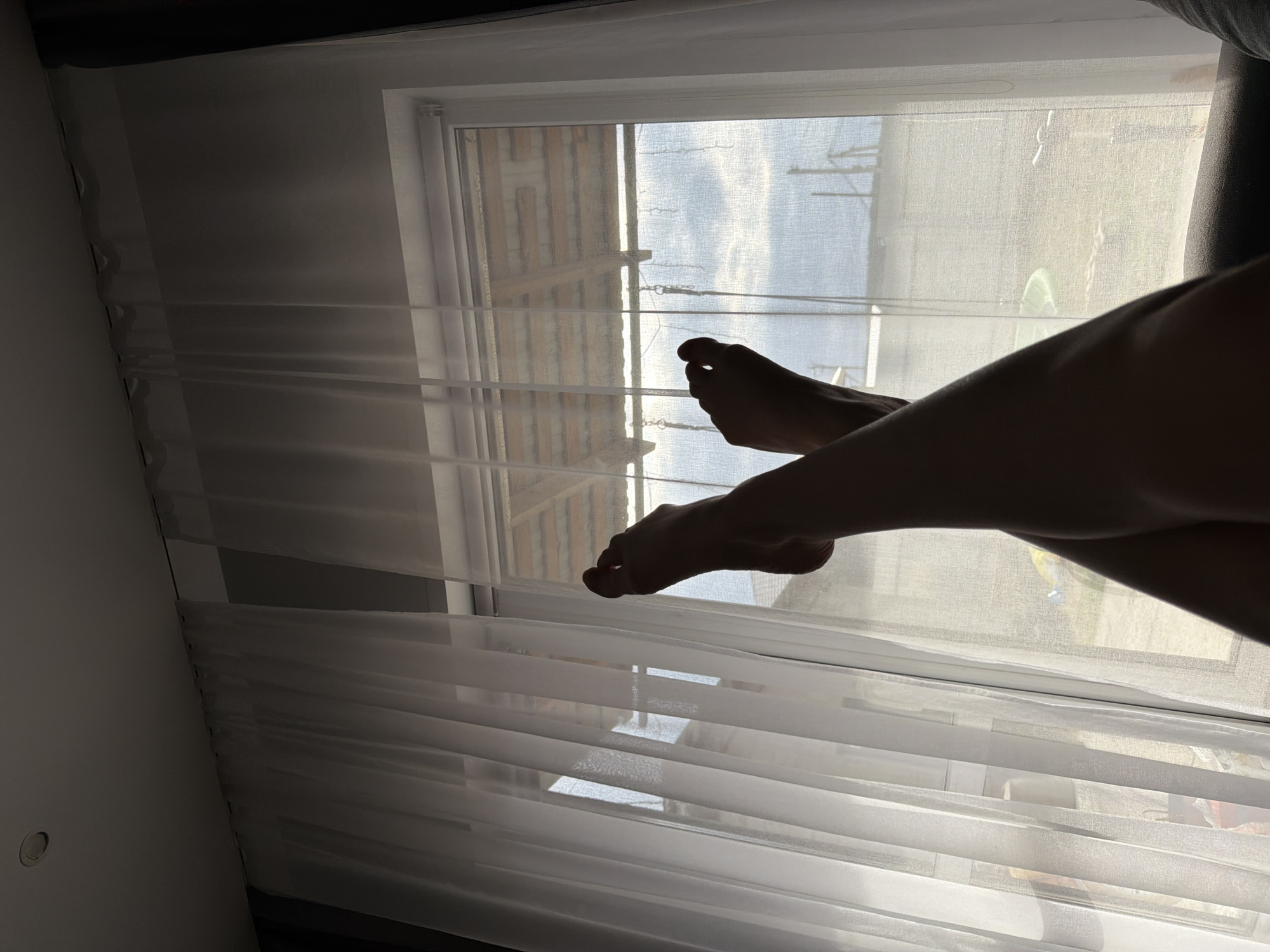 A photograph shows a pair of legs and feet silhouetted against a window covered with white, translucent curtains. Through the window, a view of a building exterior and a streetscape can be seen. The location is possibly Chaikovsky, Russia, based on the query. There are no other people visible.
