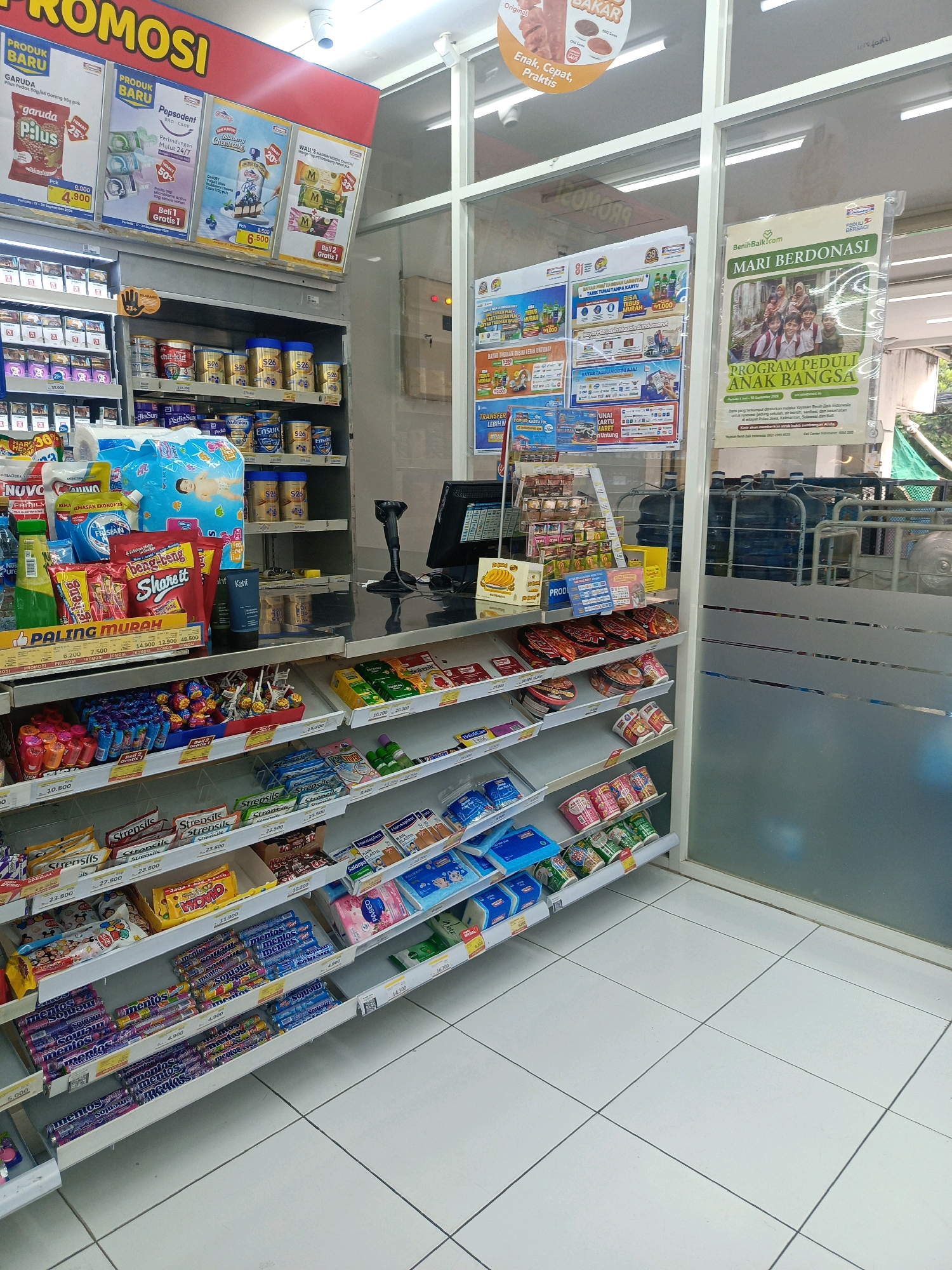 This is a photograph taken inside an Indomaret convenience store in Kepolorejo, Indonesia. The image shows a wide view of the store's interior, focusing on shelves stocked with various products. The shelves are organized and display items such as snacks, drinks, hygiene products, and baby care items. Promotional posters are visible on the walls, advertising current deals and campaigns. In the background, a checkout counter with a computer monitor and scanner is present. The floor is tiled in a white, square pattern. The overall environment appears clean and well-lit, typical of a modern convenience store. There are no people visible in the frame.