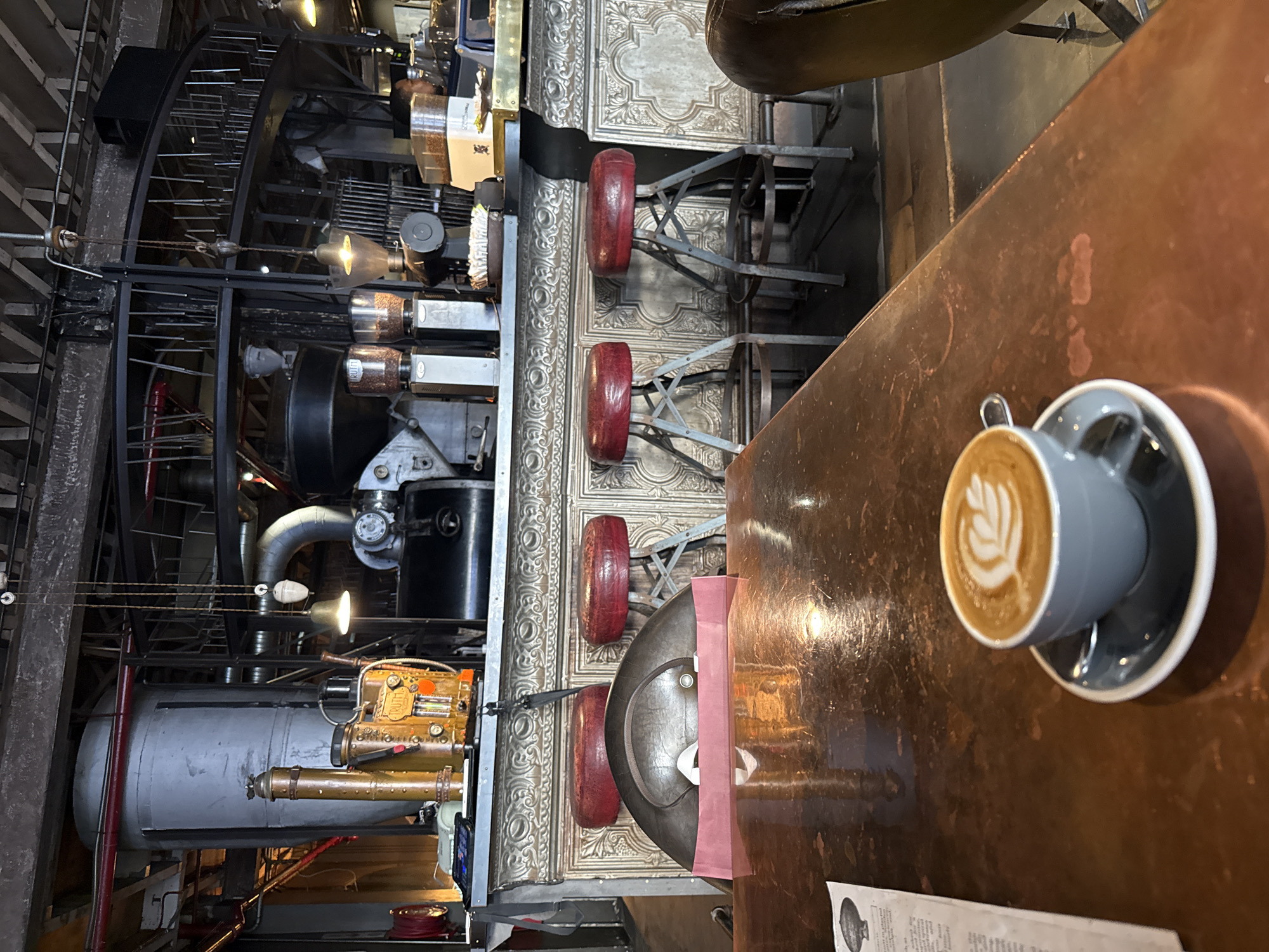 The image captures the interior of a coffee shop in Cape Town, South Africa, specifically the Truth Coffee Roasting establishment. On the right, a cup of coffee with latte art sits on a copper-colored table, next to a menu. To the left of the table, the image reveals ornate decor details, including a textured silver wall with retro-style red-seated stools. In the background, large steampunk-inspired coffee roasting machinery dominates the space, with exposed gears, pipes, and various industrial components. The overall setting appears to be an elaborately designed, industrial-chic cafe.
