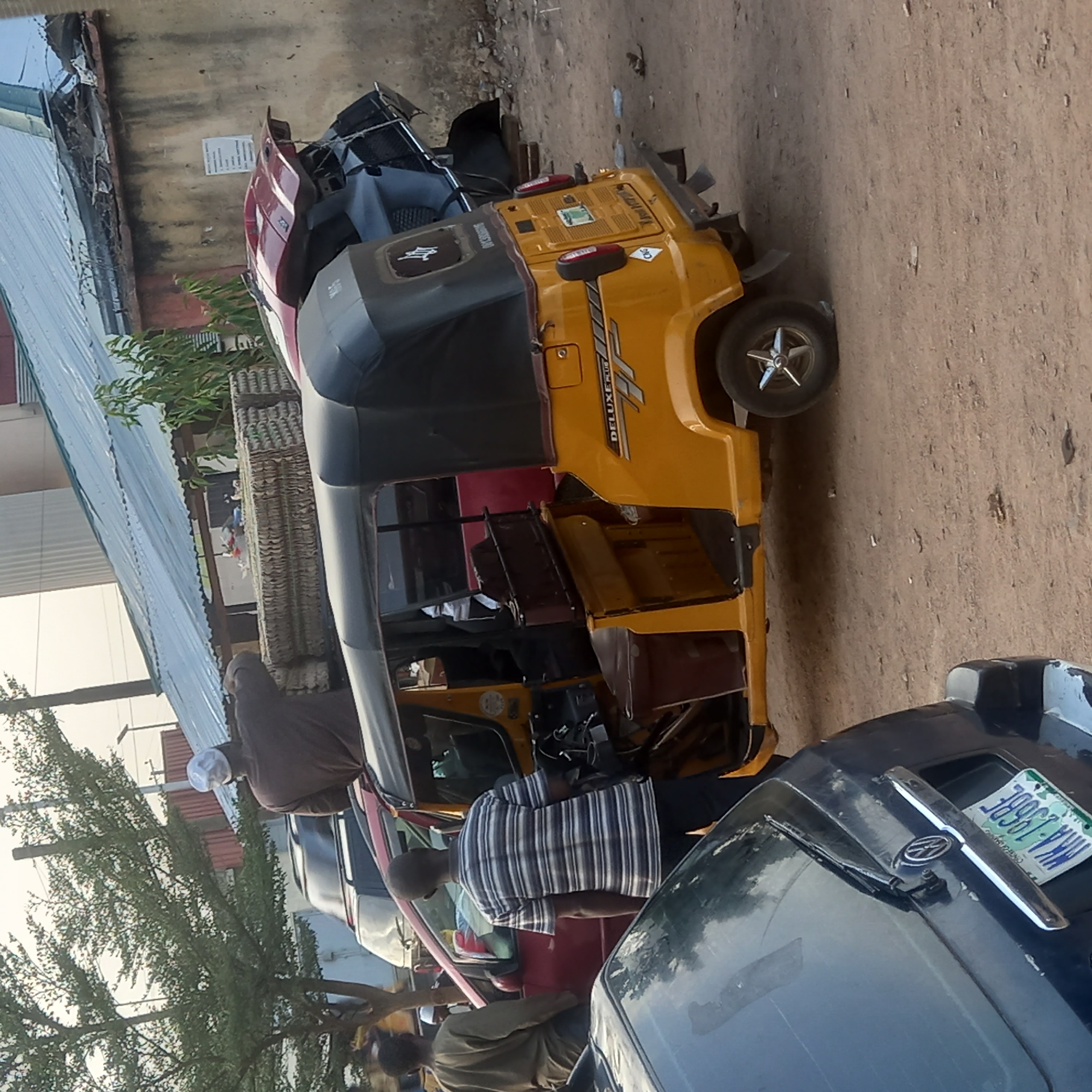 The image captures a daytime street scene in Sardauna, Nigeria, featuring several vehicles and individuals. The central focus is a yellow Bajaj RE Deluxe Plus auto-rickshaw (keke napep) parked on a dirt road, facing towards the right. The auto-rickshaw displays "DELUXE PLUS" and "BAJAJ RE" branding on its side panel, and its rear wheel is fitted with a star-shaped hubcap.
Around the auto-rickshaw, several people are visible. A man wearing a striped shirt stands beside a red car, which is partially obscured behind the auto-rickshaw. To the left, another man in a brown garment and a white cap stands near the auto-rickshaw. In the foreground on the right, a dark blue car is parked, featuring a Volkswagen logo on its bonnet and a visible license plate that reads "WKA-236BZ" with "AKWA IBOM" below it, indicating a Nigerian registration.
The background includes various buildings, some with corrugated metal roofs, and scattered trees and foliage. A light-colored wall, possibly part of a building, is visible behind the vehicles, bearing a small, unreadable white sign. The scene suggests a typical urban or semi-urban setting, with natural daylight indicating clear weather conditions. No specific activity beyond people being present near the vehicles is clearly discernible.