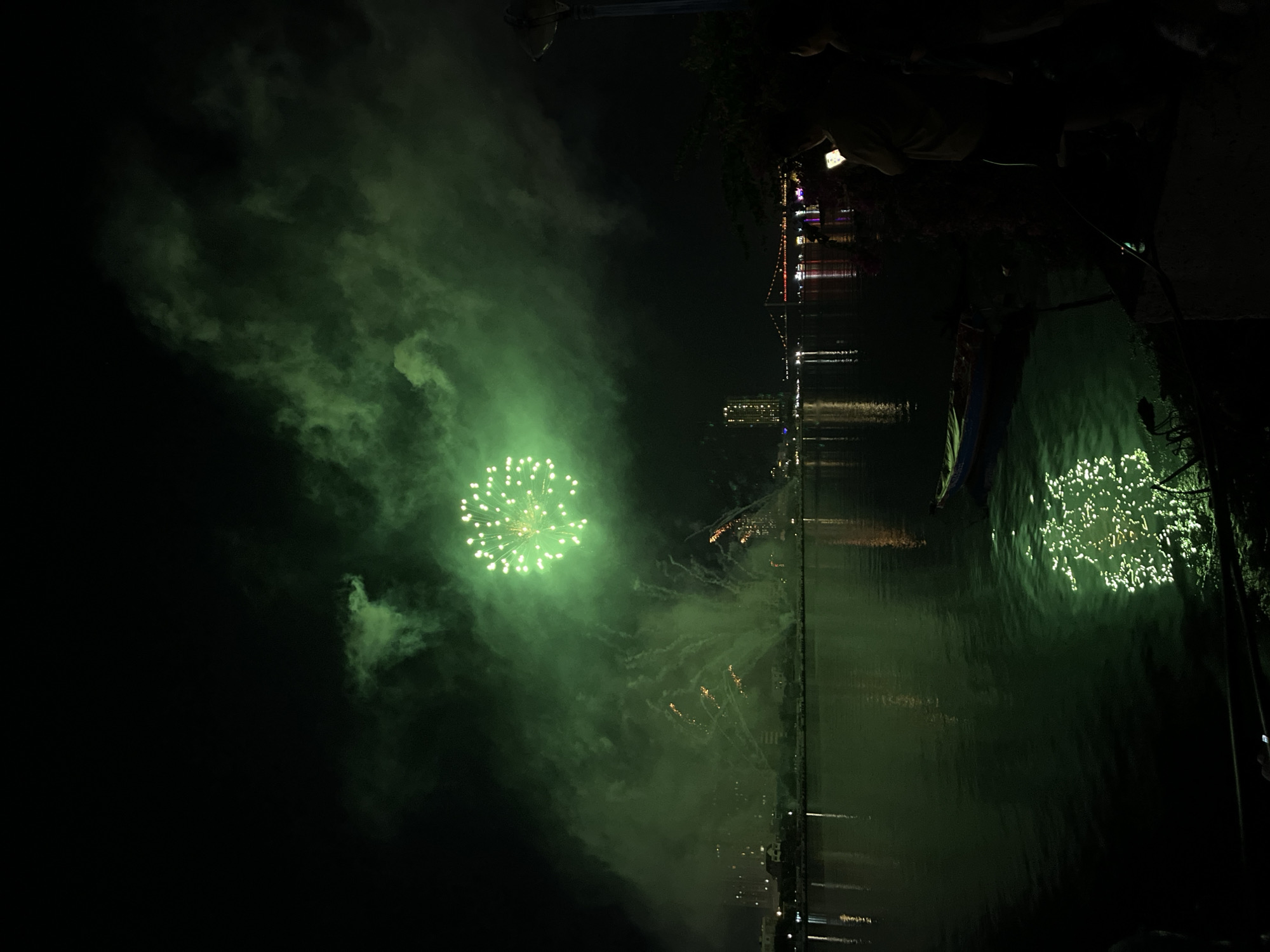 A wide-angle, low-light photograph captures a nighttime urban riverfront scene in Đà Nẵng, Vietnam, featuring a fireworks display. A large, spherical green fireworks burst is centered in the upper-middle of the frame, radiating numerous bright green points of light. This central explosion illuminates a dense cloud of green-tinged smoke that expands across the upper portion of the sky.
Reflections of the green fireworks create a sparkling pattern on the dark, rippled surface of the Hàn River in the lower-center of the image. On the far bank, the cityscape of Đà Nẵng is visible, delineated by numerous illuminated buildings and structures. The prominent Dragon Bridge, with its distinctive arch and supports illuminated in yellow-orange, spans the river in the mid-ground. Additional, smaller fireworks trails, exhibiting orange and yellow hues, ascend from the river level on both the left and right sides, contributing to the smoky atmosphere.
In the dark foreground on the right, two indistinct human figures are partially visible. One figure holds a smartphone, its bright white screen light illuminating a portion of the person's torso. A small, blue-hulled boat with a lighter-colored interior is moored close to the riverbank in the lower-right quadrant. The immediate foreground consists of a dark, uneven ground surface adjacent to the water, with some indeterminate dark structures or vegetation. A tall, dark street lamp pole, with an unlit fixture, is visible in the upper-right corner.