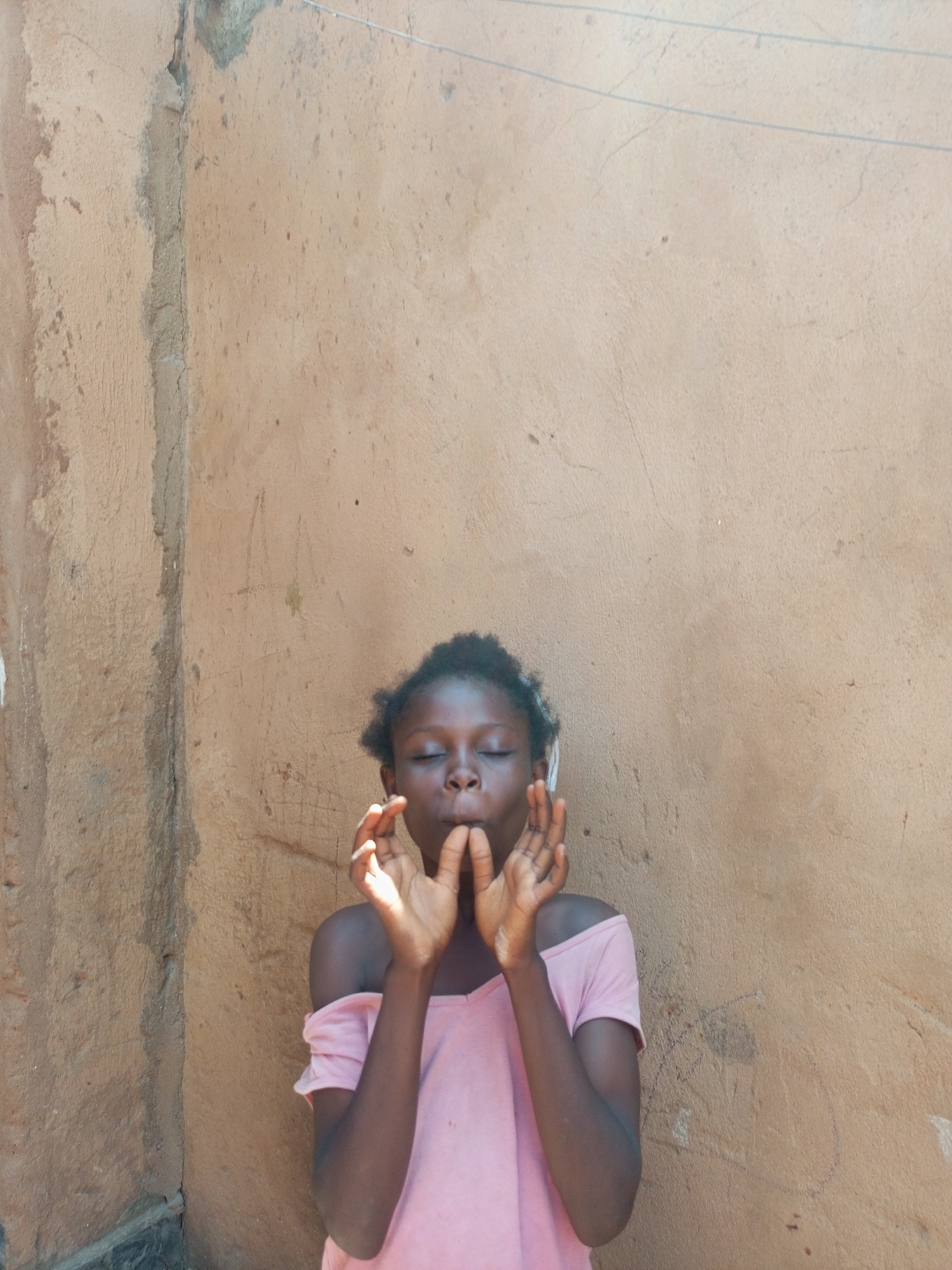 A young Black girl, wearing a light pink off-the-shoulder t-shirt, stands in front of a textured, light brown wall. Her eyes are closed, and her hands are held up to her mouth, as if she is whistling or making a sound. Her dark, curly hair is short and messy. The wall appears to be an exterior wall of a building, with some cracks and signs of wear. The lighting suggests it is daytime. The location is identified as a street in Tempure, Nigeria.