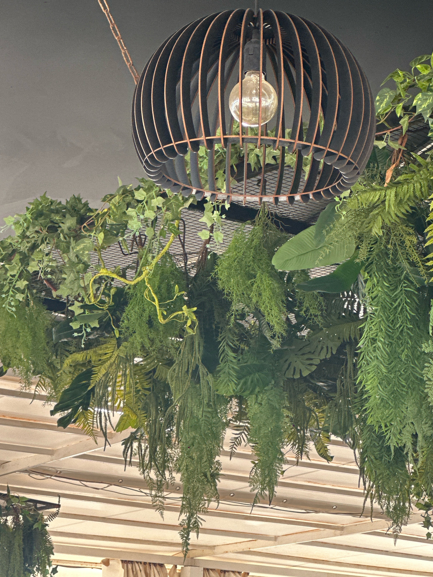 This image depicts a close-up view of a decorative hanging light fixture with a birdcage-like design, suspended from a chain. The light fixture is surrounded by an abundance of lush, artificial greenery, including ferns and ivy, creating a jungle-like atmosphere. The greenery hangs down from what appears to be a mesh structure above, cascading around the light. Below the greenery, a white slatted ceiling or awning is visible. The overall scene suggests an indoor or covered outdoor space, possibly a restaurant or cafe, with a bohemian or naturalistic decor. There are no people or animals in the image. The location is identified as Lermontovo, Russia.