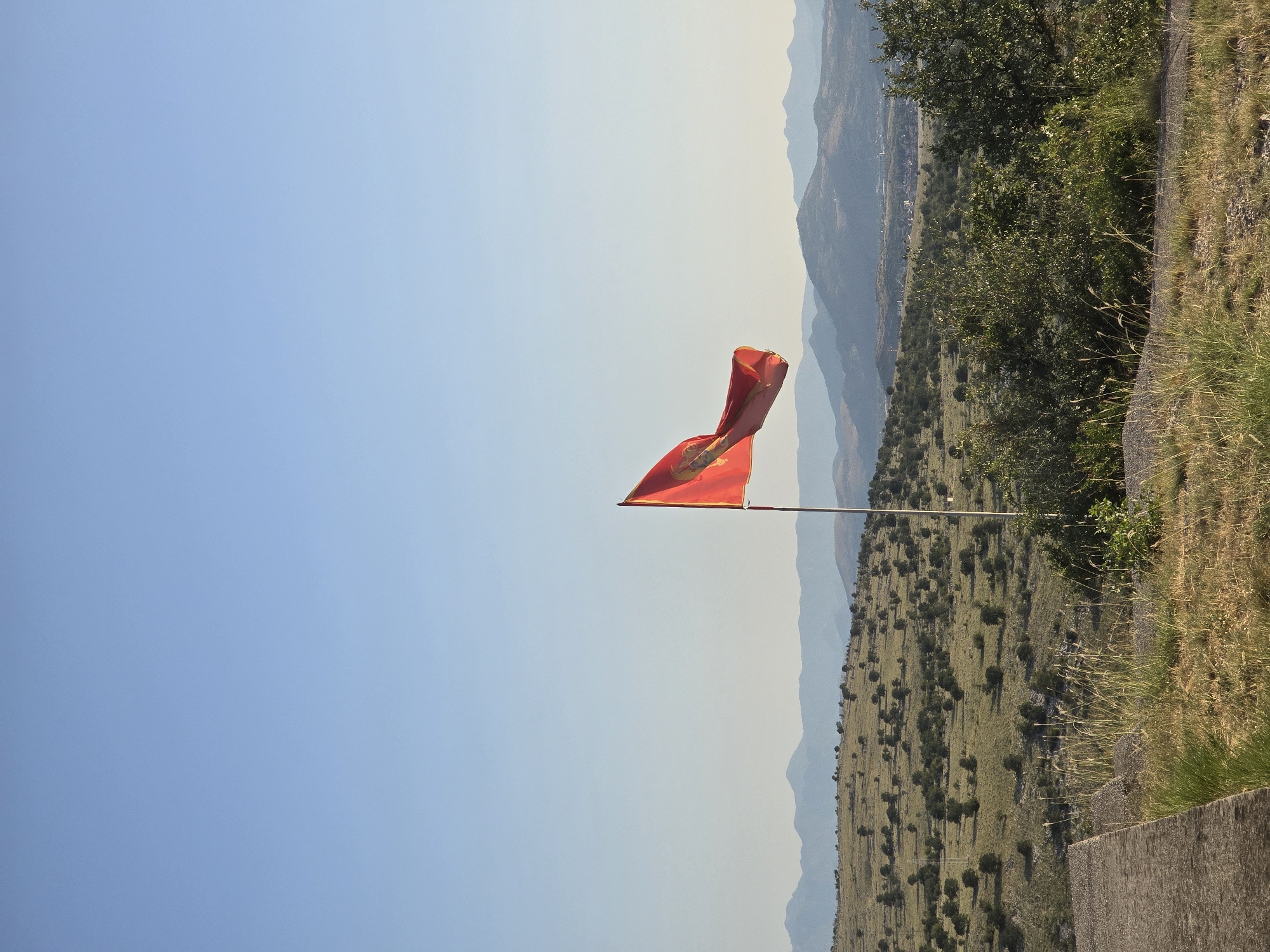 A vibrant red flag with a golden emblem is prominently displayed on a flagpole against a clear, pale blue sky. The flag is positioned to the left of the frame, its fabric rippling in the breeze. In the background, a landscape of rolling hills unfolds, dotted with scattered green trees and scrub. Further in the distance, a range of hazy, blue-toned mountains stretches across the horizon. The foreground features dry, golden grass and some greenery, with a section of a concrete structure visible on the lower left. The scene appears to be an outdoor, elevated viewpoint overlooking a rural or mountainous area.