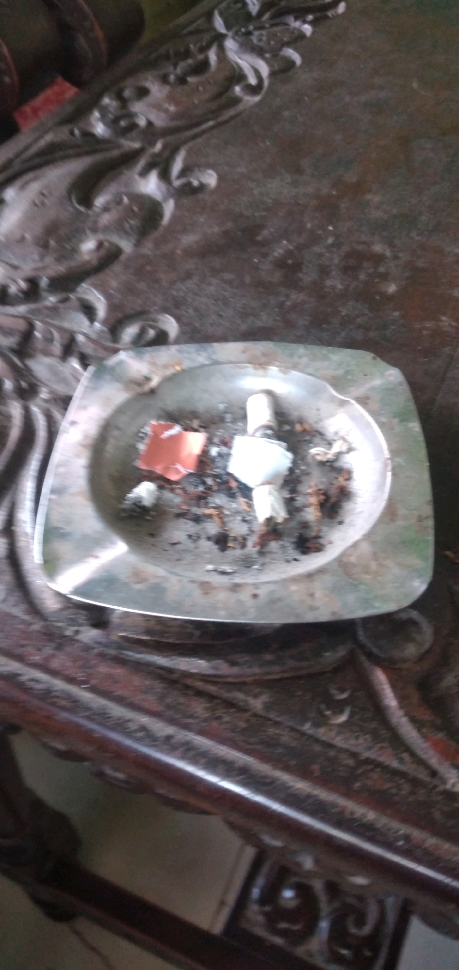 A high-angle, slightly blurry, close-up shot shows a silver ashtray filled with cigarette butts, ashes, and a small piece of orange paper. The ashtray is on a dark, ornate wooden table. The table appears to be in an indoor setting. There is no indication of the specific location within Rahong, Indonesia, beyond it being a general Indonesian locale.