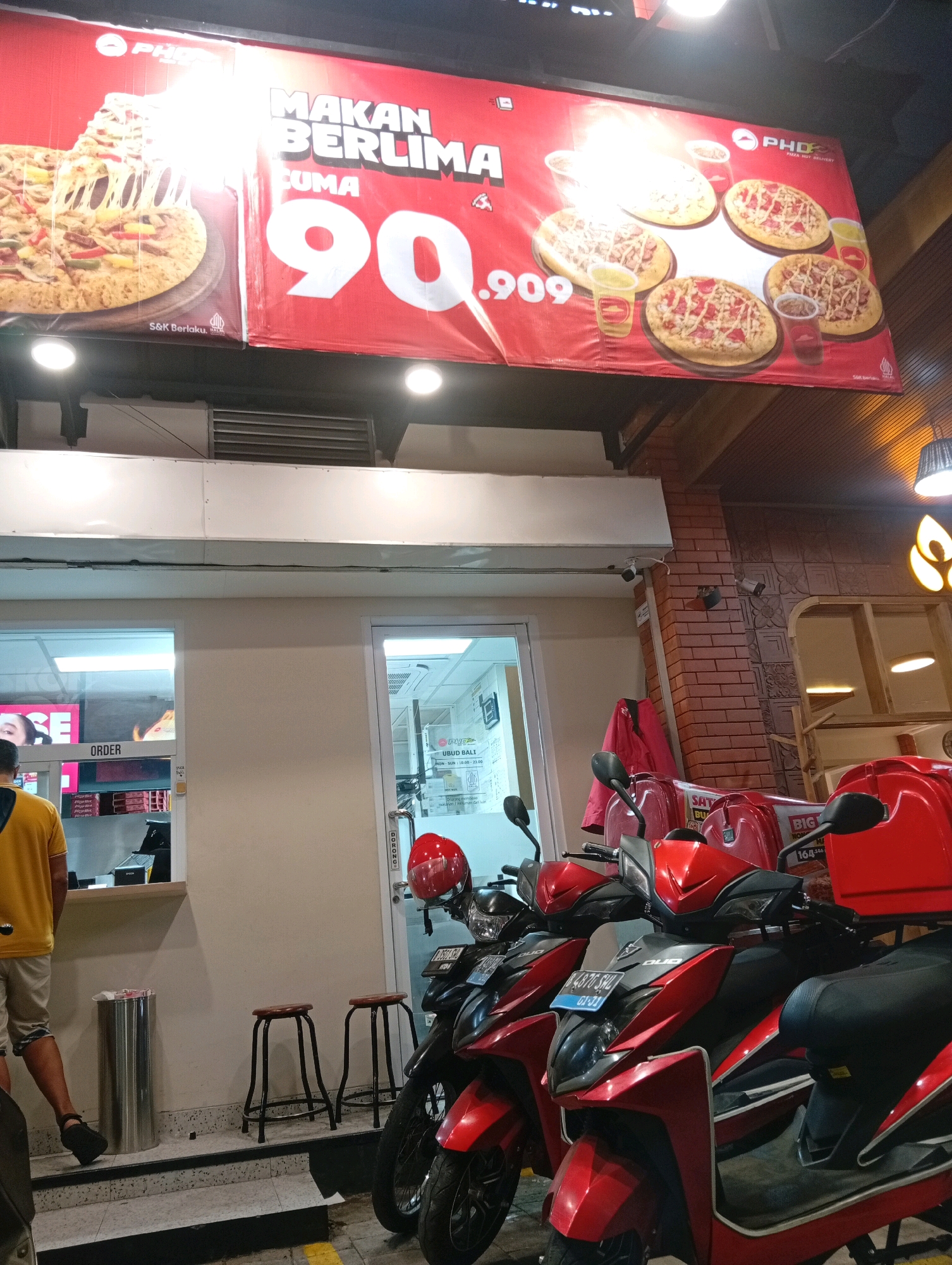 An outdoor promotional banner for PHD Pizza Hut Delivery is displayed above an entrance to a food establishment in Ubud District, Indonesia. The banner features a large image of a pizza with melting cheese and text advertising a "Makan Berlima Cuma 90.909" promotion, meaning "Eat Five for Only 90,909." Several smaller images of pizzas and drinks are also present on the banner.
Below the banner, a person wearing a yellow shirt and shorts is visible from the back, standing near a counter with an "ORDER" sign. To the right, a red door with a "DORONG" (push) sign is observed. Several red and black scooters are parked in front of the establishment, some equipped with red delivery boxes. Two small stools are placed near the entrance. The environment appears to be a commercial area with brick and tiled walls and overhead lighting.