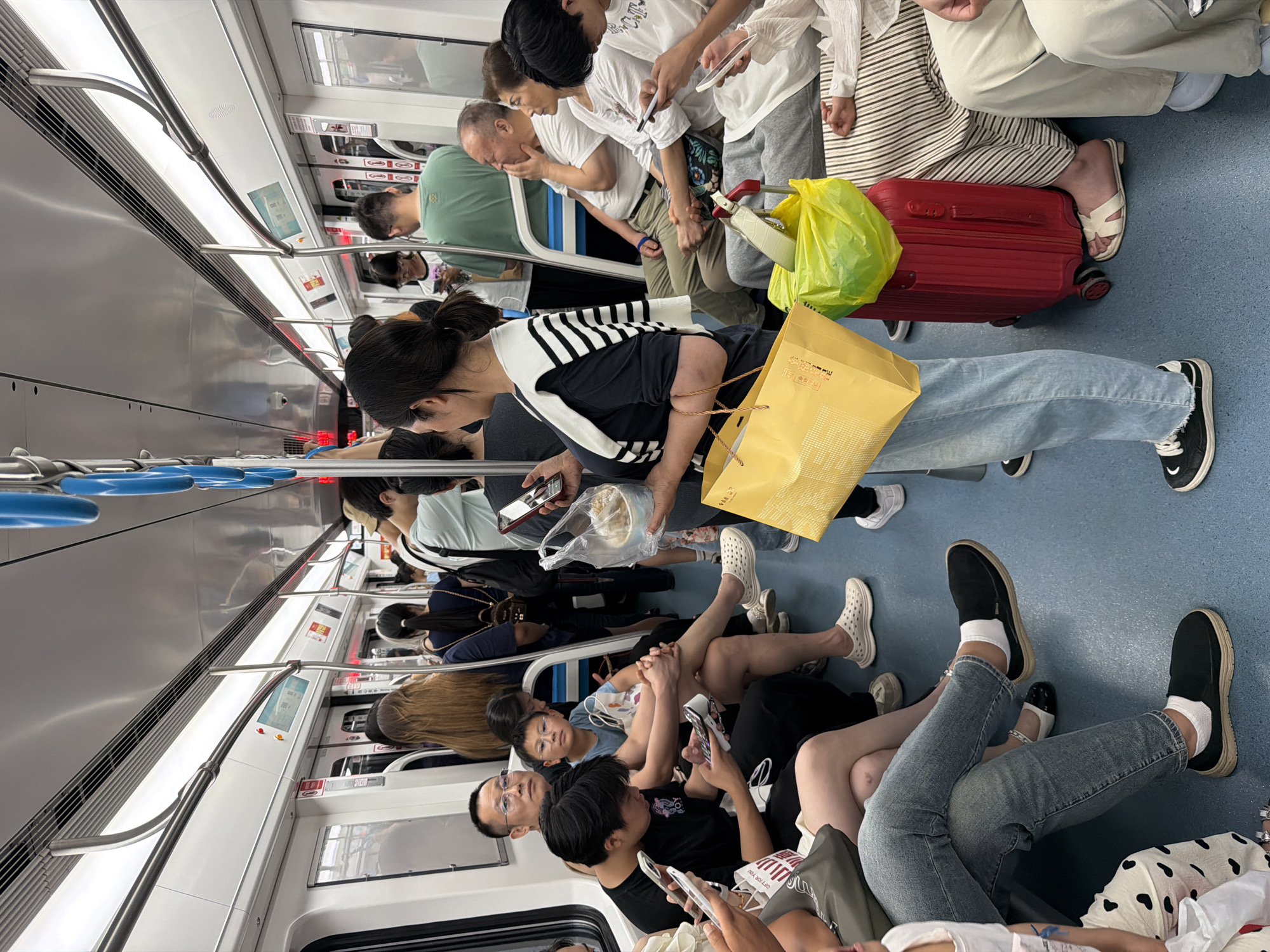 This image captures the interior of a subway train in Unknown, Unknown, occupied by numerous passengers. The train car features white walls, a light blue floor, overhead fluorescent lighting, silver horizontal and vertical handrails, and blue hanging grab handles. Windows are visible along the sides of the car.
On the left side of the image, multiple individuals are seated. In the foreground, a person wearing light-wash jeans, white socks, and black slip-on shoes is seated with legs crossed. Next to them, a person with short dark hair, wearing a black t-shirt with "BOY" text, is engrossed in a smartphone. Behind them, a man with glasses and a light-colored polo shirt is seated, looking forward. Adjacent to him, a young boy with glasses and a blue t-shirt is also seated, looking to the left. Other partially visible passengers are seated or standing further back on this side.
On the right side, a woman with dark hair tied in a ponytail stands prominently in the middle ground. She wears a black t-shirt with a white and black striped sweater draped over her shoulders. She holds a yellow shopping bag in her left hand and a clear plastic bag containing food in her right hand, while looking at a smartphone. At her feet, a red hard-shell rolling suitcase is positioned on the floor, topped with a bright green plastic bag and a light-colored belt. Seated behind the suitcase are several passengers, including an older man in a white polo shirt with his hand resting on his chin, an older woman in a white t-shirt next to him, and another individual in a white t-shirt and striped pants, using a smartphone. A foot in a white sandal is visible.
Throughout the car, many passengers, both seated and standing, are engaged with their smartphones. The train appears moderately crowded. The environment is clean and well-lit, characteristic of a modern public transport system. Red and white signage, along with digital displays, are visible above the doors and along the upper walls, but the specific text or images are not discernible.