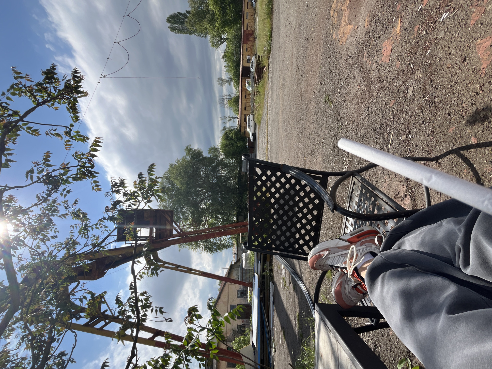 The image depicts a person sitting on a black metal park bench outdoors. The person is wearing grey sweatpants and white and orange sneakers. A white poster tube rests on the bench next to them. The setting appears to be an industrial area in Burgas, Bulgaria, as evidenced by the presence of an old, rusting crane structure, and dilapidated buildings. The ground is covered in gravel. Trees are visible, obscuring a view of the sky with cloudy weather. A powerline is also present.