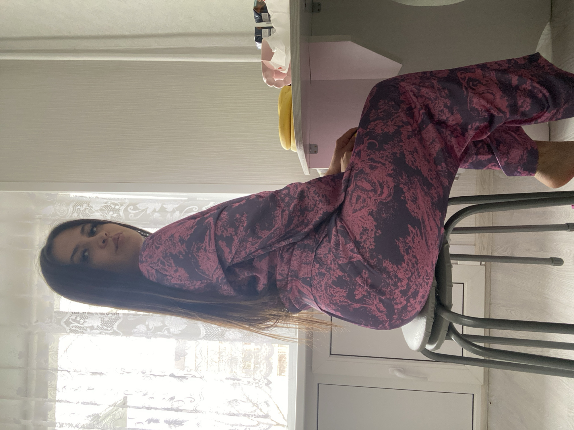 A woman is seated on a metal stool indoors, wearing purple pajamas with a pink pattern. She is positioned near a window covered with a lace curtain. The background reveals a desk with various items on it. The location is suggested to be in the city of Chaikovsky, Russia.