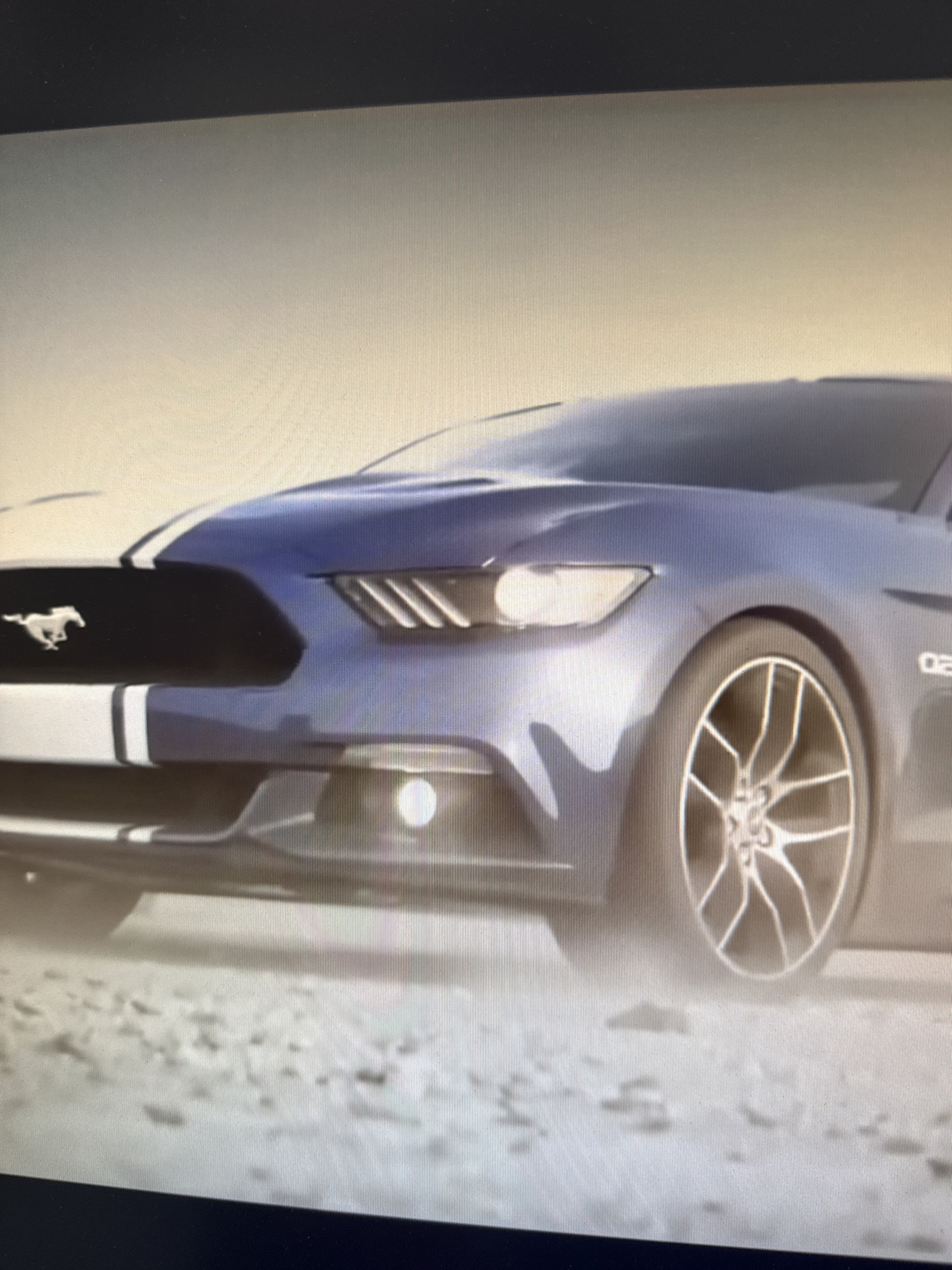 The image shows a close-up, angled view of the front of a blue Ford Mustang. The car has white racing stripes on the hood and a white Mustang emblem. The headlights are on, and there is a slight fog or dust effect around the car. The car is on a surface that appears to be dirt or gravel. The background is a hazy, light-colored sky. There are no people visible in the image. The image does not contain any information that would allow for the identification of its location in Netanya, Israel.
