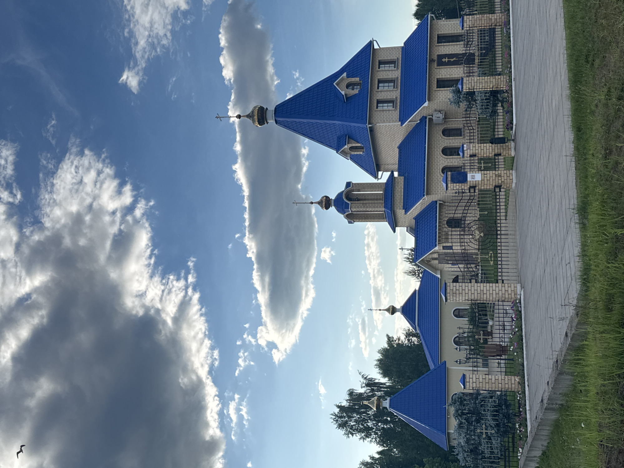 This image depicts an Eastern Orthodox church complex located in the city of Chaikovsky, Russia. The central structure is a multi-story building constructed of light-colored brick, featuring a prominent, steep, conical roof of bright blue metal sheeting, topped with a golden onion dome and an Orthodox cross. To the left, a smaller bell tower-like structure also presents a blue roof and a golden onion dome with a cross. Further to the left, a detached single-story building has a blue pyramidal roof crowned with another golden onion dome and cross. Additional structures with matching blue roofs are visible to the right, one featuring a large door adorned with an Orthodox cross.
The complex is enclosed by a black metal fence with alternating brick pillars. The fence incorporates decorative elements and ornate gates in the center, flanked by brick pillars with integrated lighting fixtures. Within the fenced perimeter, the ground is paved, and landscaped areas contain various shrubs, including a distinctive blue spruce near the main gate, and flowerbeds. Outside the fence in the foreground, there is a concrete curb separating the paved area from an unmaintained grassy strip. Deciduous and coniferous trees are visible in the background, suggesting a semi-rural or suburban setting. The sky above is a mix of deep blue and large, white-edged grey clouds, indicating direct sunlight from the right. A single dark bird is captured in flight in the upper left corner of the frame, silhouetted against a large cloud. No human figures are present in the scene.