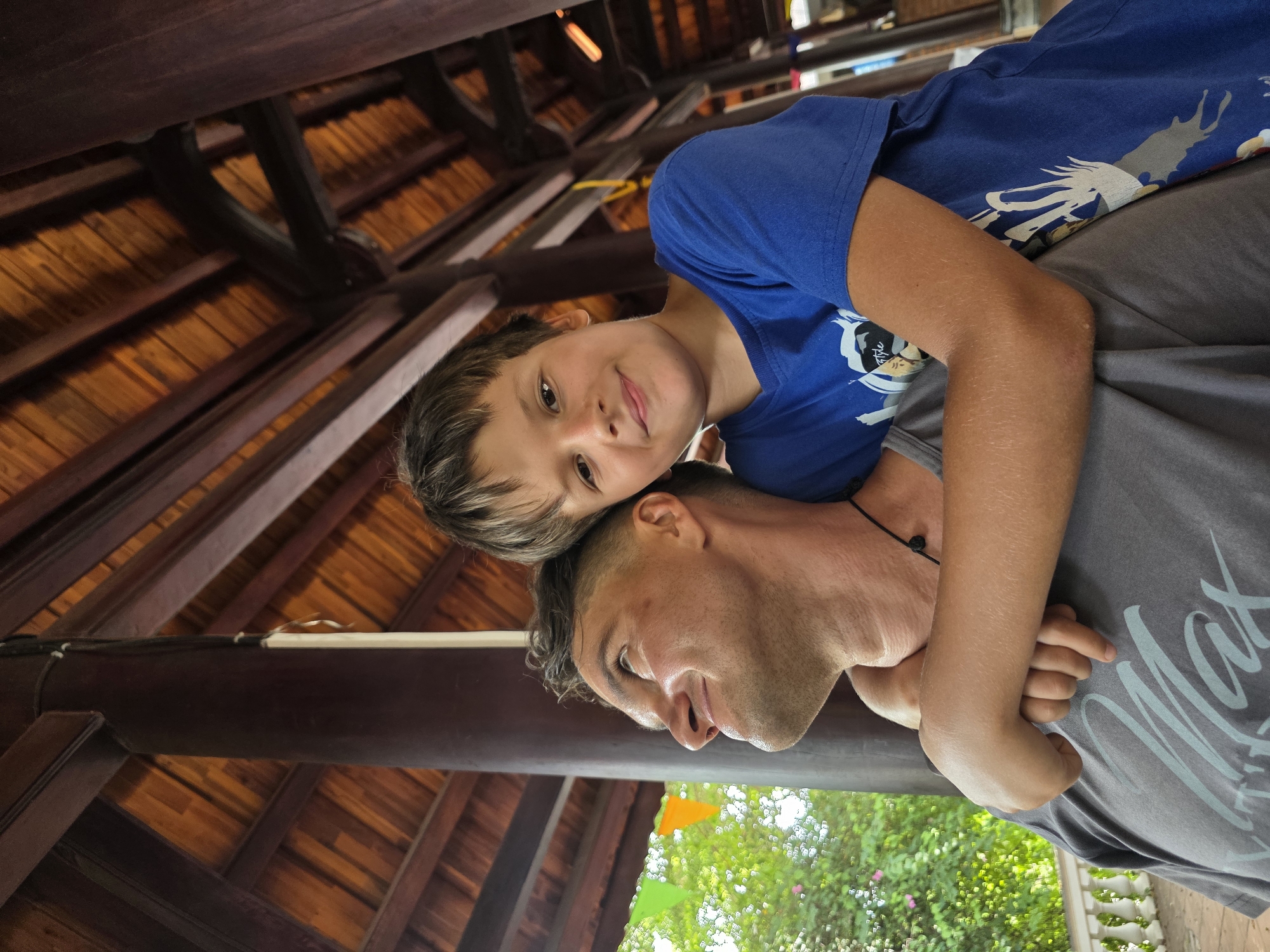 In Mui Ne, Vietnam, a young boy with short, brown hair and a blue t-shirt hugs a man from behind. The boy is smiling, looking directly at the camera, with his arms wrapped around the man's chest. The man, wearing a grey t-shirt, is looking up and to his left with a smile. They are positioned under a wooden-beamed roof, likely part of an outdoor pavilion or covered walkway. Green foliage and small, colorful triangular flags are visible in the background to the left, suggesting an outdoor setting with some decorative elements. The wooden structure has dark brown, thick pillars and a ceiling constructed from wooden planks. The lighting suggests it is daytime.