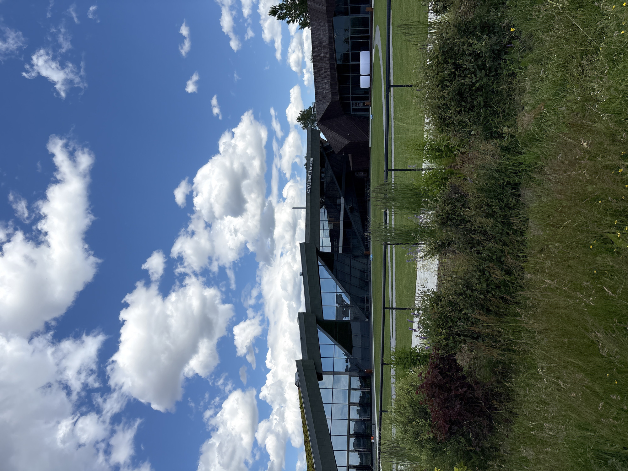 The image depicts a modern building complex on a sunny day with a blue sky and scattered white clouds. The building features a unique architectural design with sharp angles, large glass windows, and a green roof. The exterior is a combination of dark-colored materials, including what appears to be wood paneling on a section of the building and metal or glass panels on the main structure. The building is situated in an urban or suburban setting, with a well-maintained lawn in front and a black fence separating it from a foreground area with lush vegetation, including tall grasses and various shrubs. In the background, a sign reads "ROYAL BEACH РЕСТОРАН" suggesting a restaurant or event venue. Two white rectangular structures are visible on the lawn. The overall impression is one of contemporary design integrated with a natural environment. The location is the Royal Beach complex in Saint Petersburg, Russia.