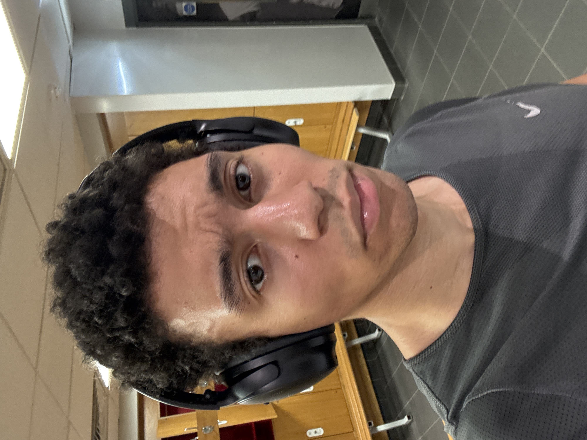 A close-up shot of a man with curly black hair and headphones. He is wearing a dark grey athletic shirt. His skin appears slightly sweaty. The background shows tiled floors and light-colored cabinets, possibly in a gym or locker room setting. The location is likely not in London, United Kingdom due to the lack of identifying features.